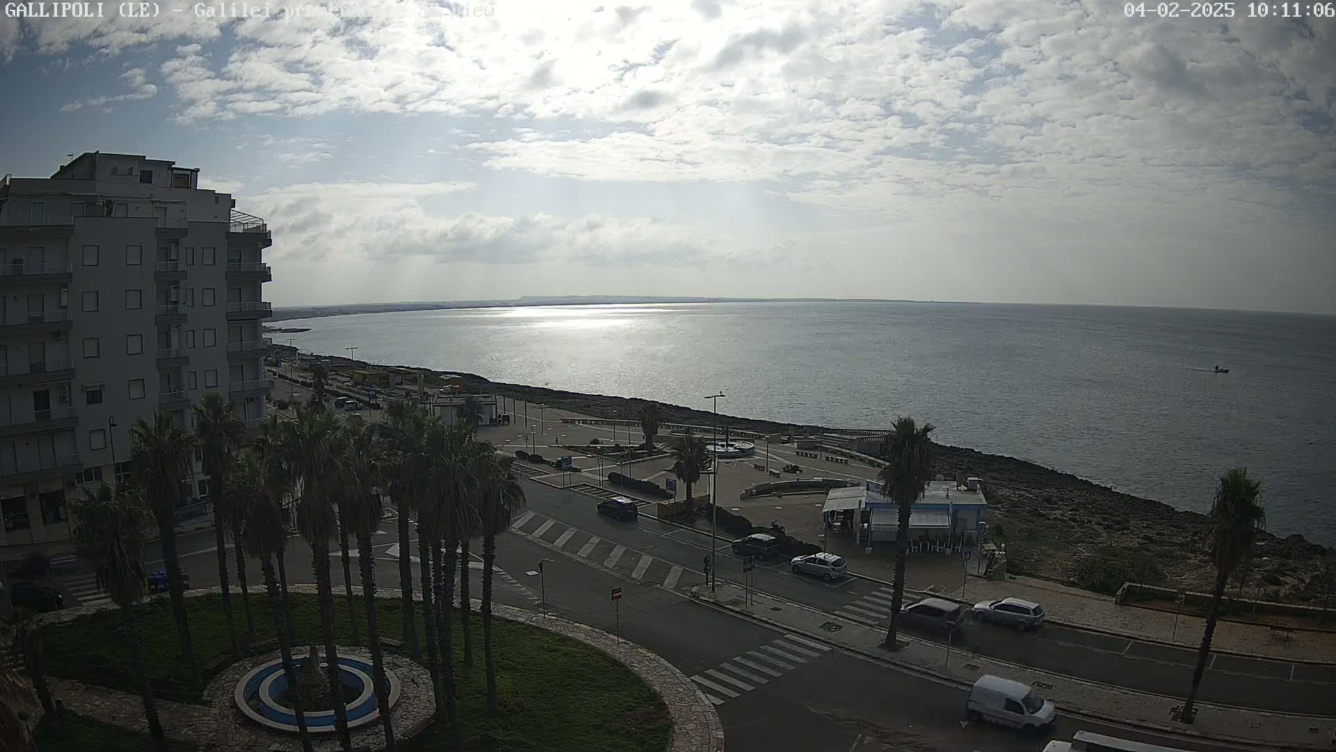 A partly cloudy day reveals a coastal town scene with a calm sea, a multi-story building, palm trees lining a roadway, and a few cars parked along the street.