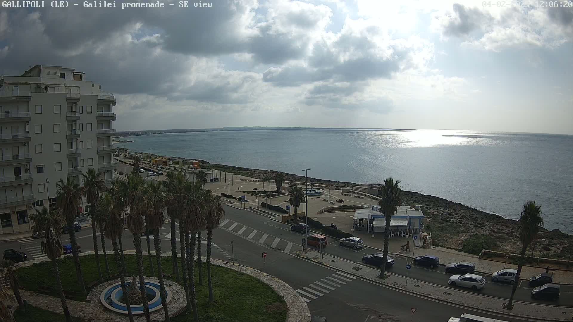 A partly cloudy day reveals a seaside town with palm trees lining a street overlooking a calm sea.