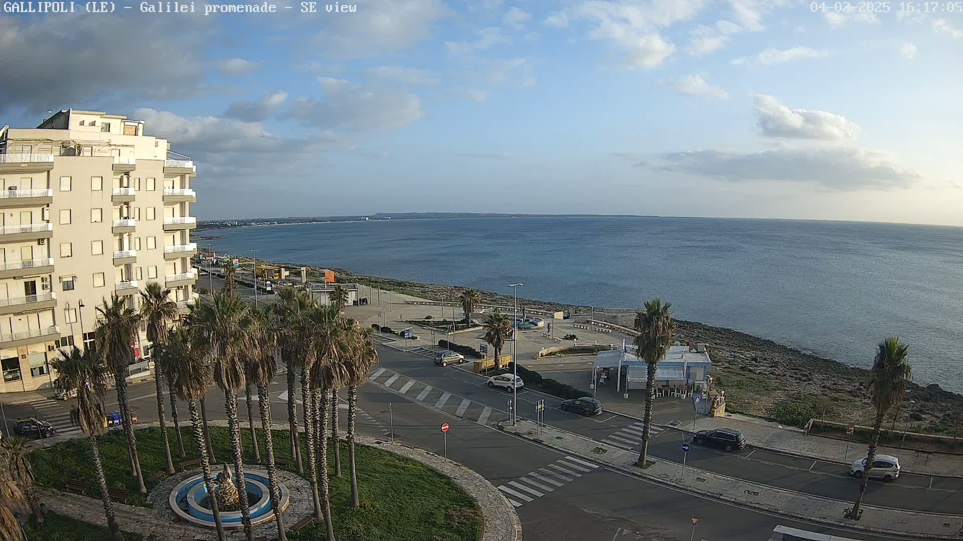 A seaside town with palm trees, a multi-story building, and cars parked along streets overlooking a calm sea under a partly cloudy sky.