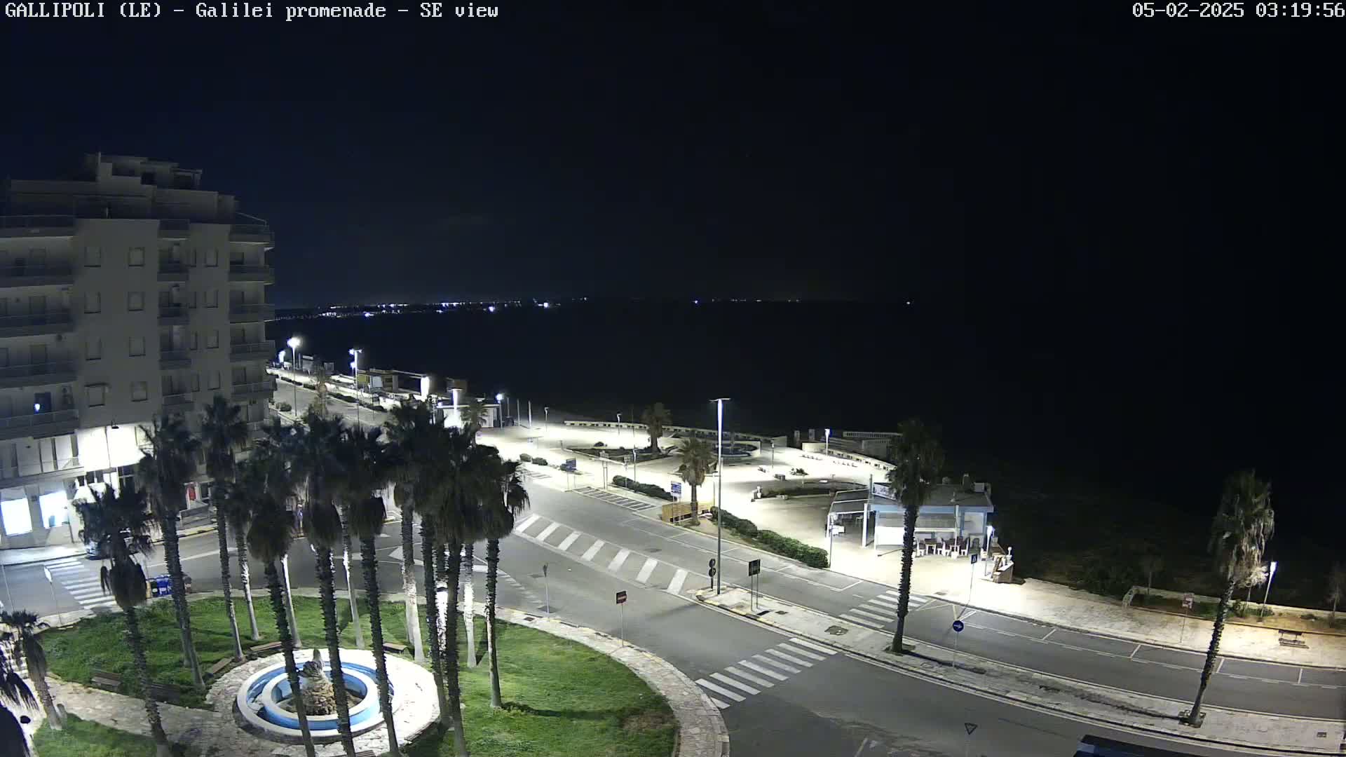 A nighttime, high-angle view shows a seaside promenade with palm trees, a fountain, and a multi-story building, overlooking a dark body of water.