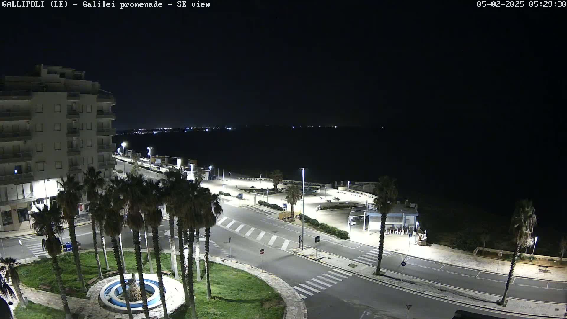 A nighttime, high-angle view of a coastal town square with palm trees surrounding a fountain, a multi-story building, and a dark body of water in the background.