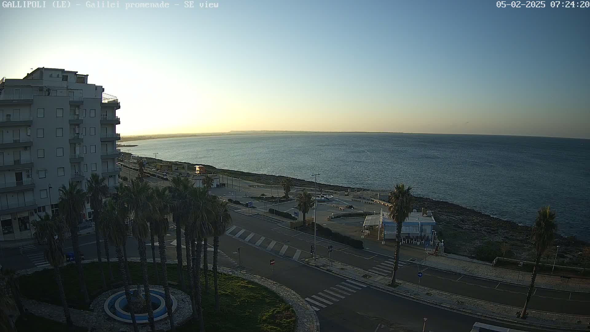 A clear sunrise illuminates a coastal town with a multi-story building, palm trees lining a promenade overlooking a calm sea.