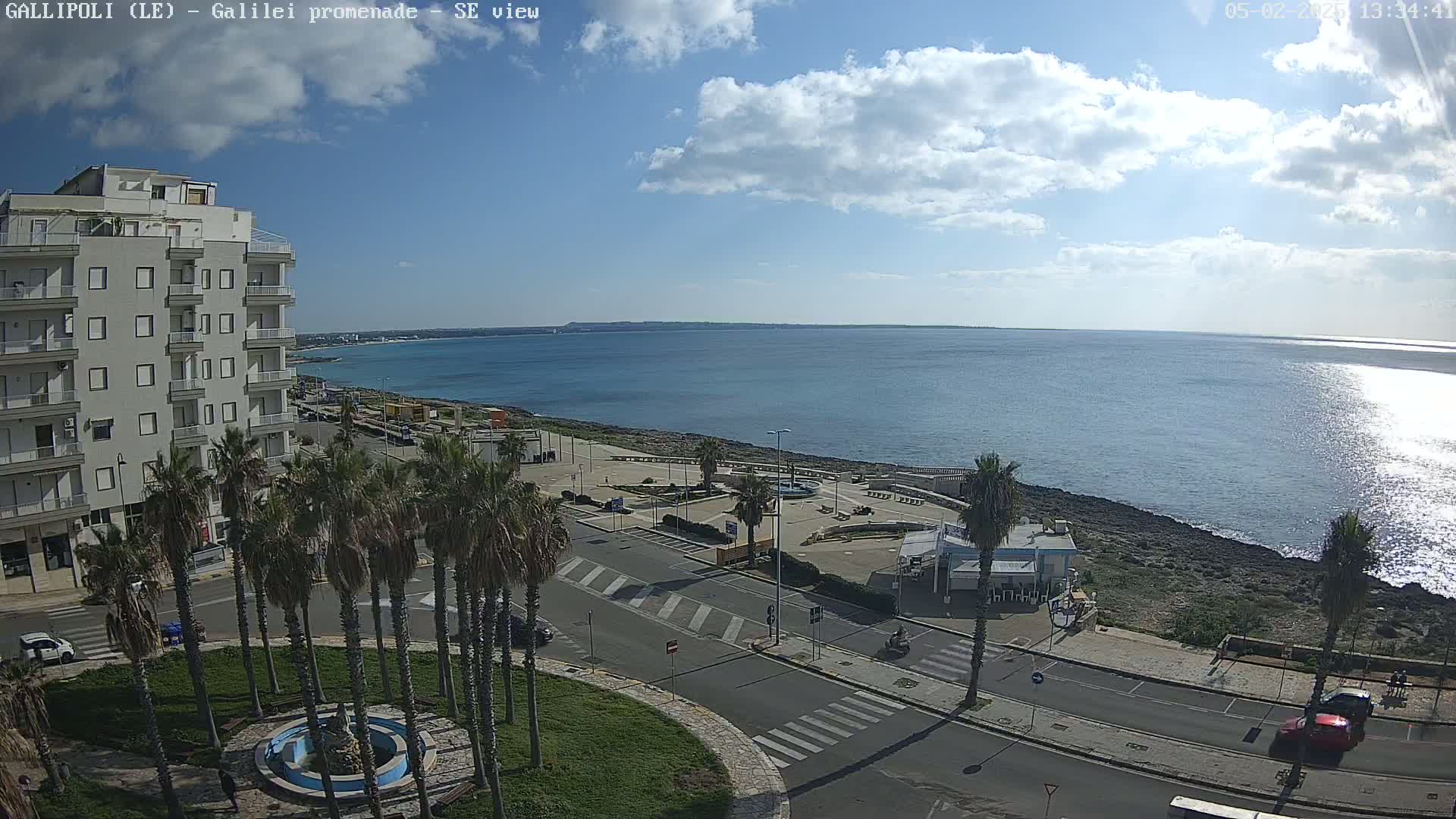 A partly cloudy day shows a coastal town scene with a calm sea, palm trees lining a street next to a rocky shoreline, and a multi-story building in the background.
