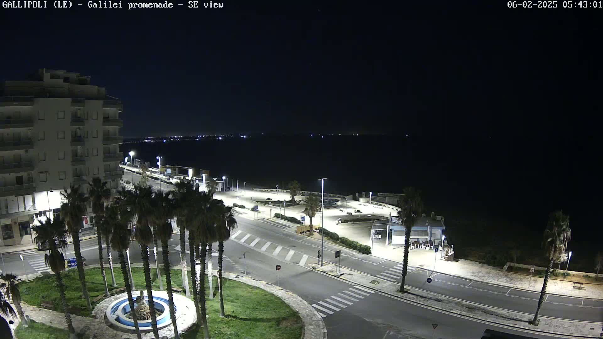 A nighttime view of a seaside promenade with palm trees, a fountain, and a multi-story building, under a clear, dark sky.