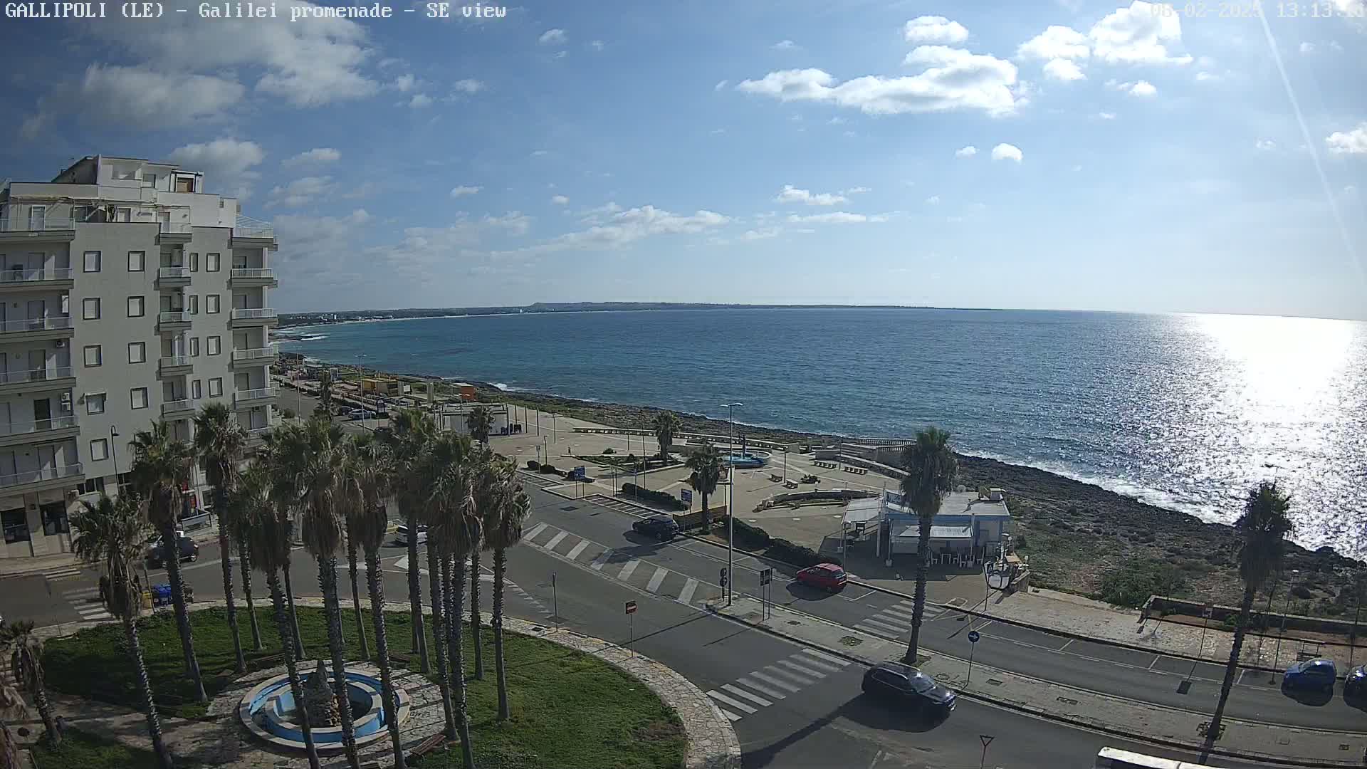 A partly sunny day reveals a seaside town with a promenade, palm trees, a multi-story building, and several vehicles parked along the road, overlooking a calm ocean with some ripples reflecting sunlight.