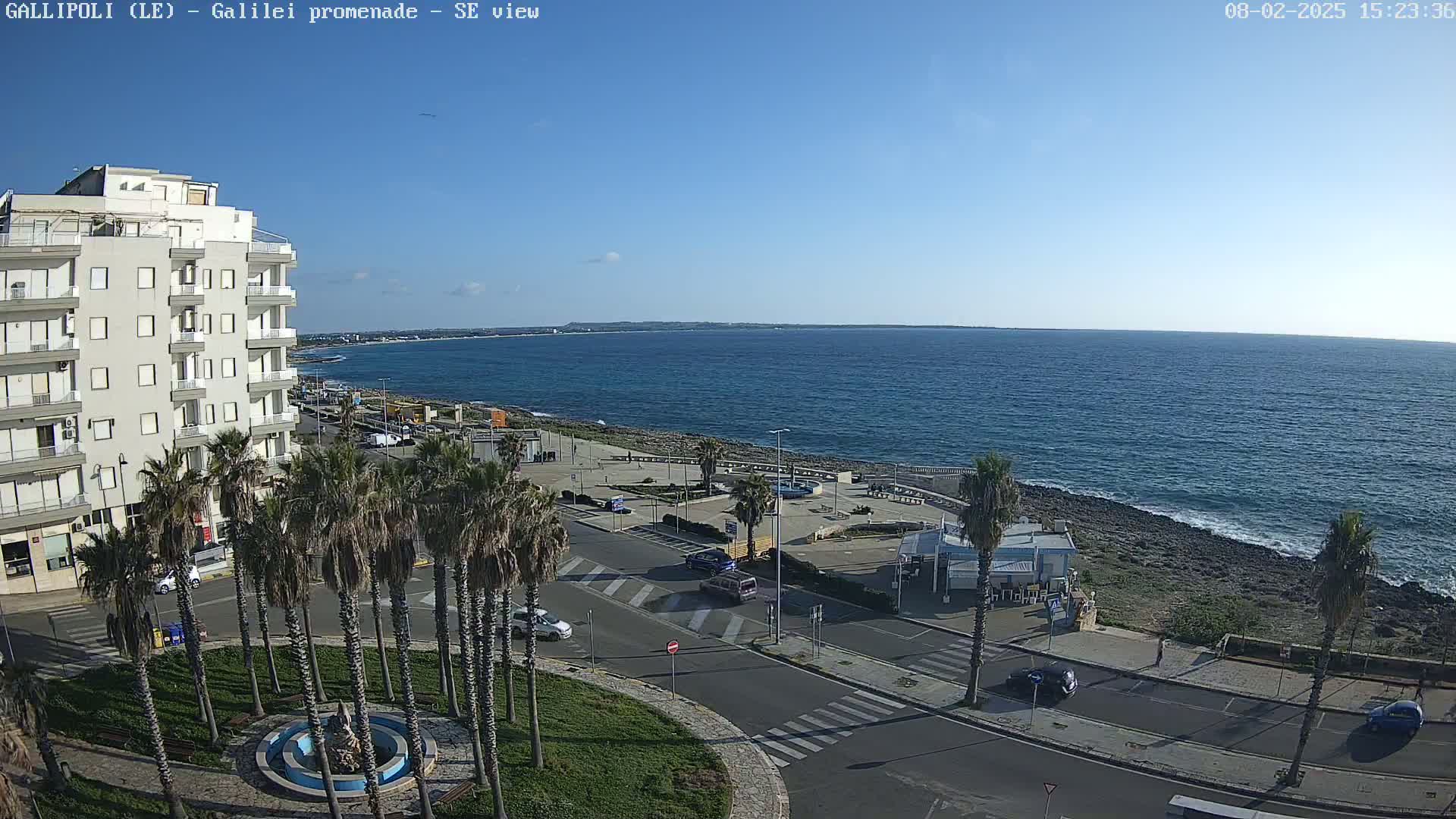 A seaside town scene on a sunny day shows a multi-story building, palm trees lining a street near the ocean, and a rocky shoreline with calm, blue water extending to the horizon.