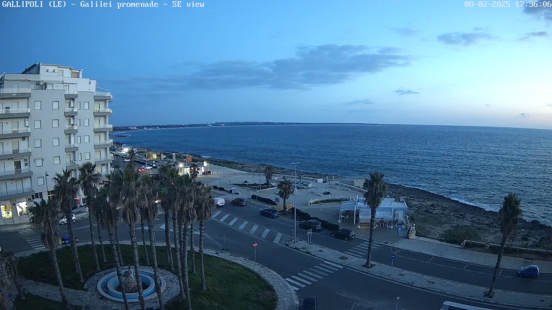 A seaside town at dusk, featuring a multi-story building, palm trees lining a road next to a promenade, and a calm sea under a mostly clear sky.