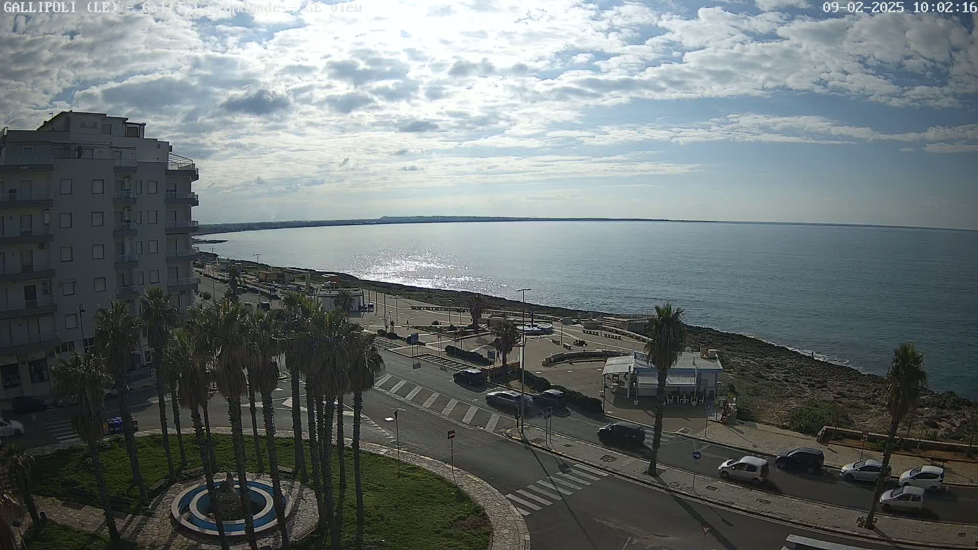 A partly cloudy day overlooks a coastal town with a calm sea, palm trees lining a street next to a building and a seaside promenade.