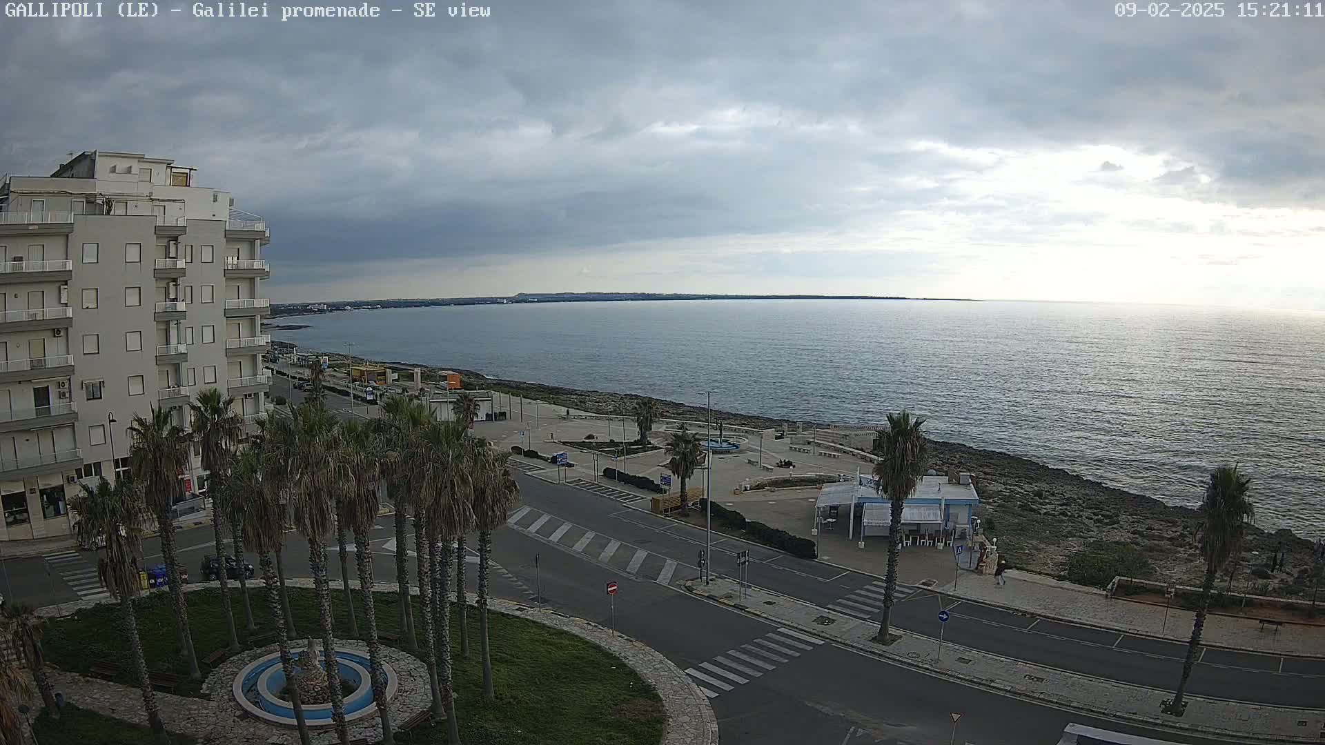 An overcast day shows a coastal town scene with a multi-story building, palm trees lining a promenade next to a calm sea.