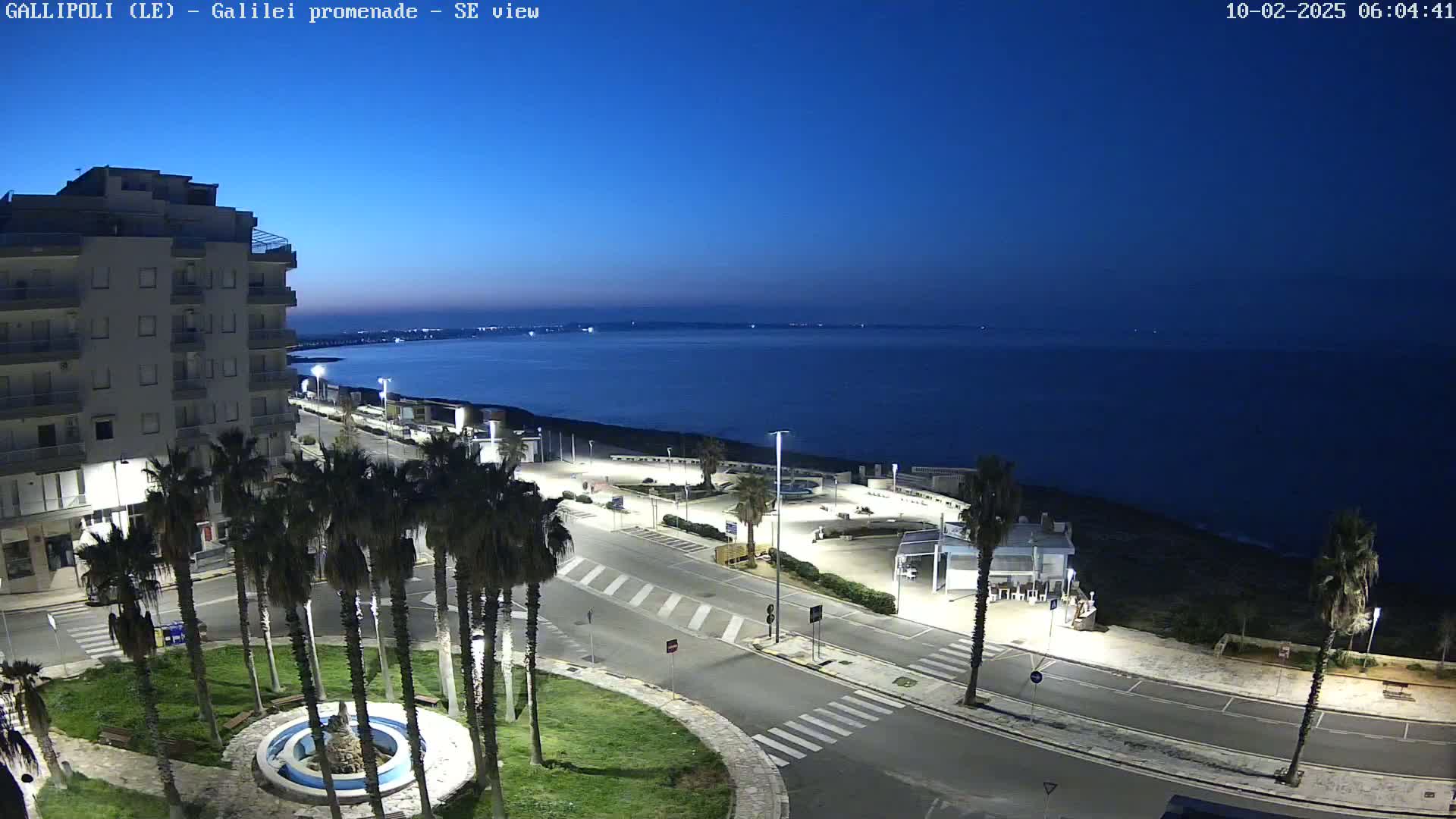 A coastal town scene at twilight shows a multi-story building, palm trees lining a roadway near the ocean, and a calm, dark-blue sea under a clear, deep-blue sky.