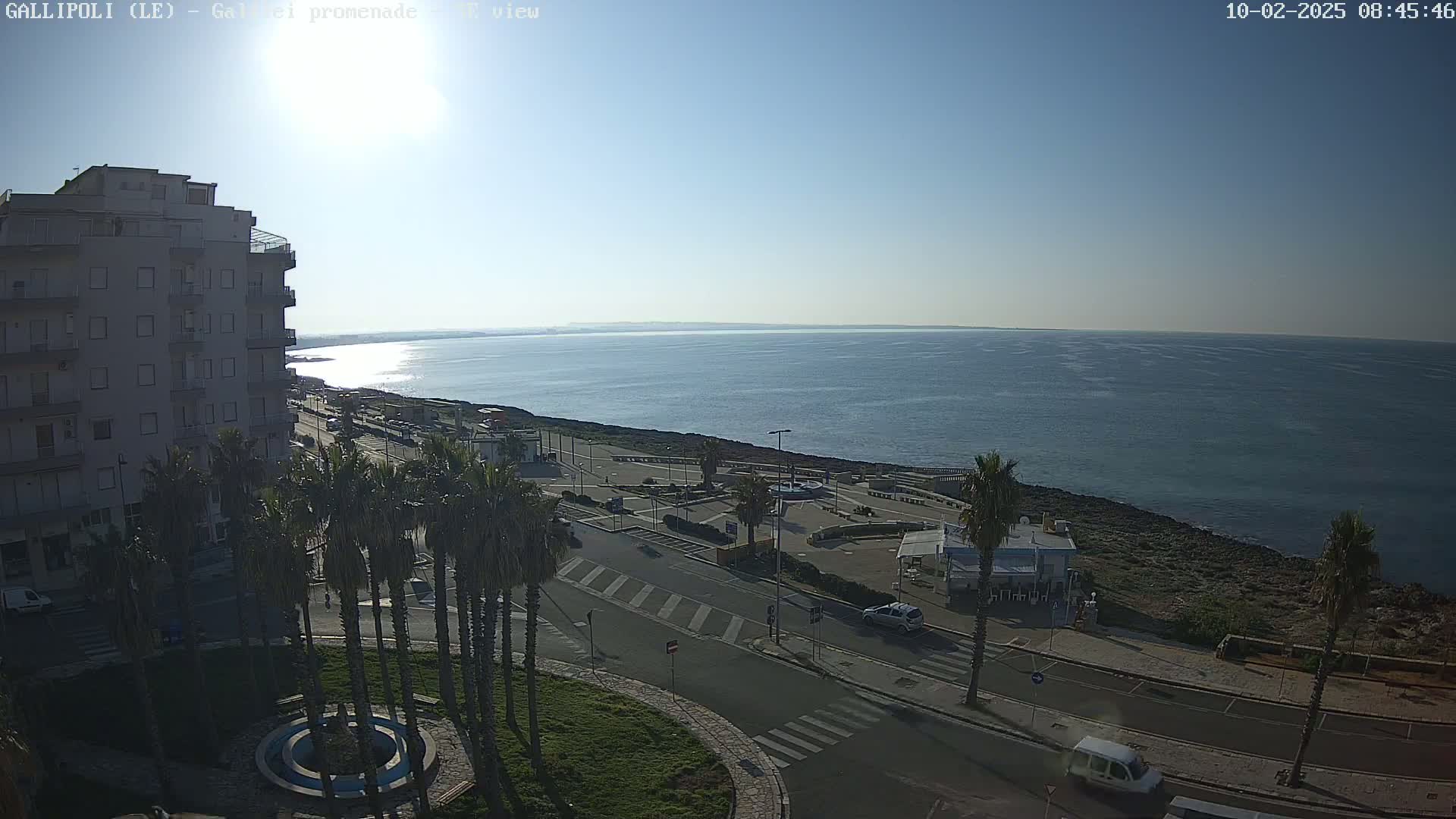 A sunny day overlooks a calm sea from a high vantage point, showing a coastal town with palm trees, buildings, and a roadway leading to a seaside plaza.