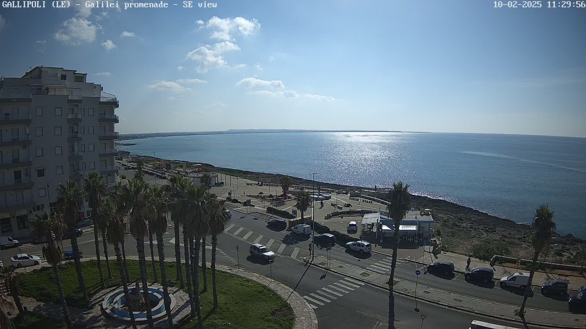 A mostly sunny day overlooks a coastal town with a promenade, palm trees, buildings, and parked cars, all next to a calm, sparkling blue sea.