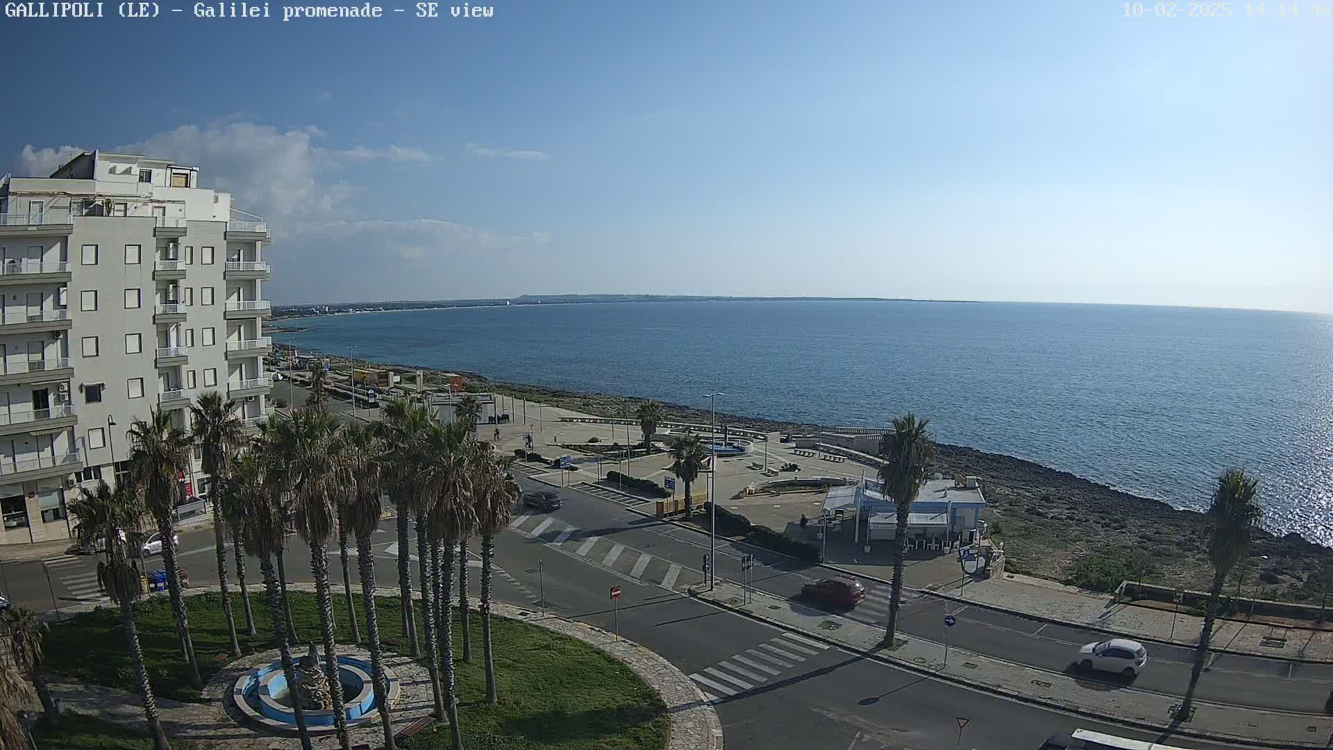 A seaside town scene on a partly cloudy day shows a promenade with palm trees, cars, and a building overlooking a calm sea.