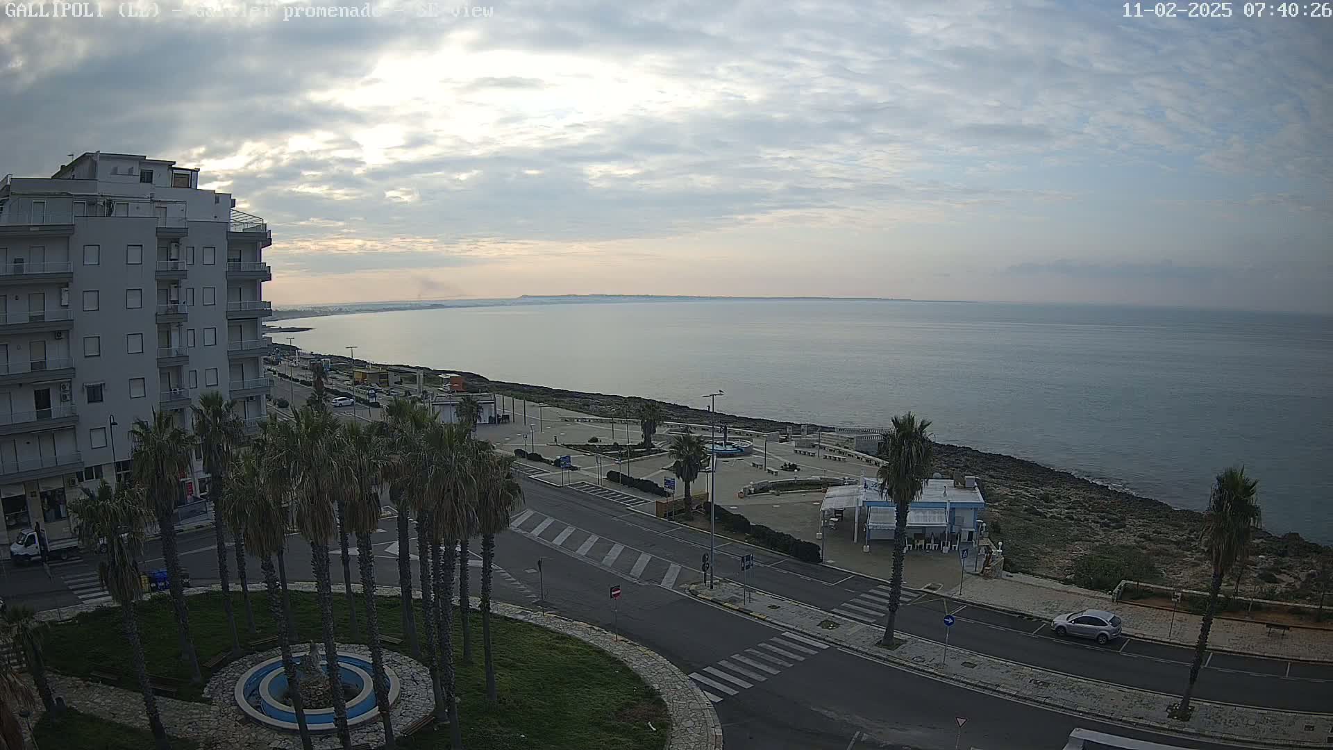 A mostly cloudy sunrise overlooks a calm sea from a seaside town with palm trees lining a street next to a promenade.