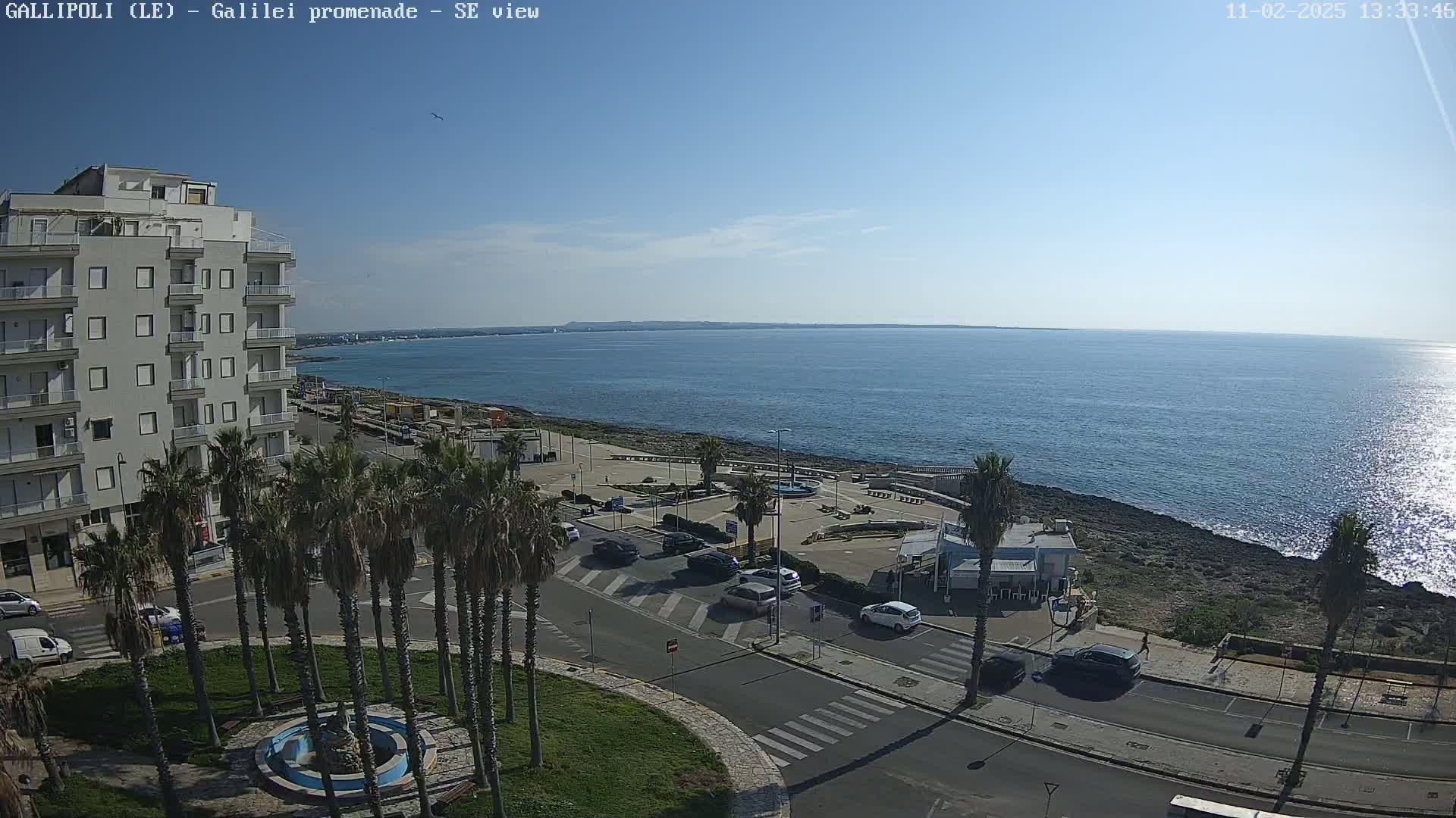 A coastal town scene on a sunny day shows a multi-story building, palm trees, cars on a road near a waterfront promenade with a calm sea extending to the horizon.