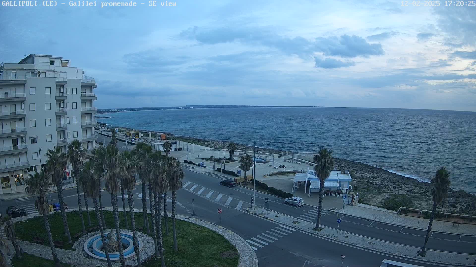 A partly cloudy day overlooks a coastal road lined with palm trees, leading to a calm sea with a distant shoreline.