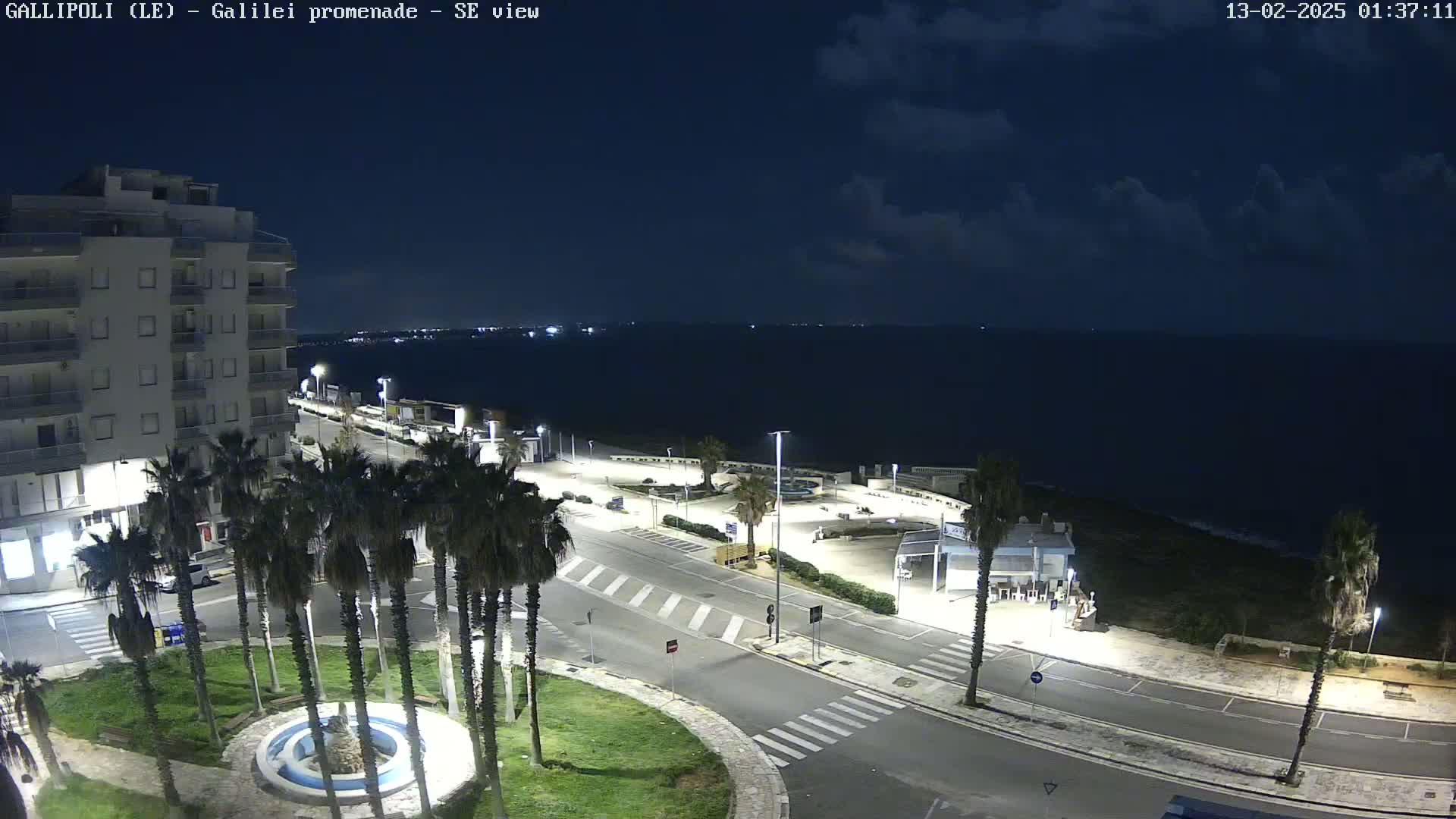 A nighttime, overhead view of a coastal town shows a mostly empty promenade, a multi-story building, palm trees, and a calm sea under a partly cloudy sky.