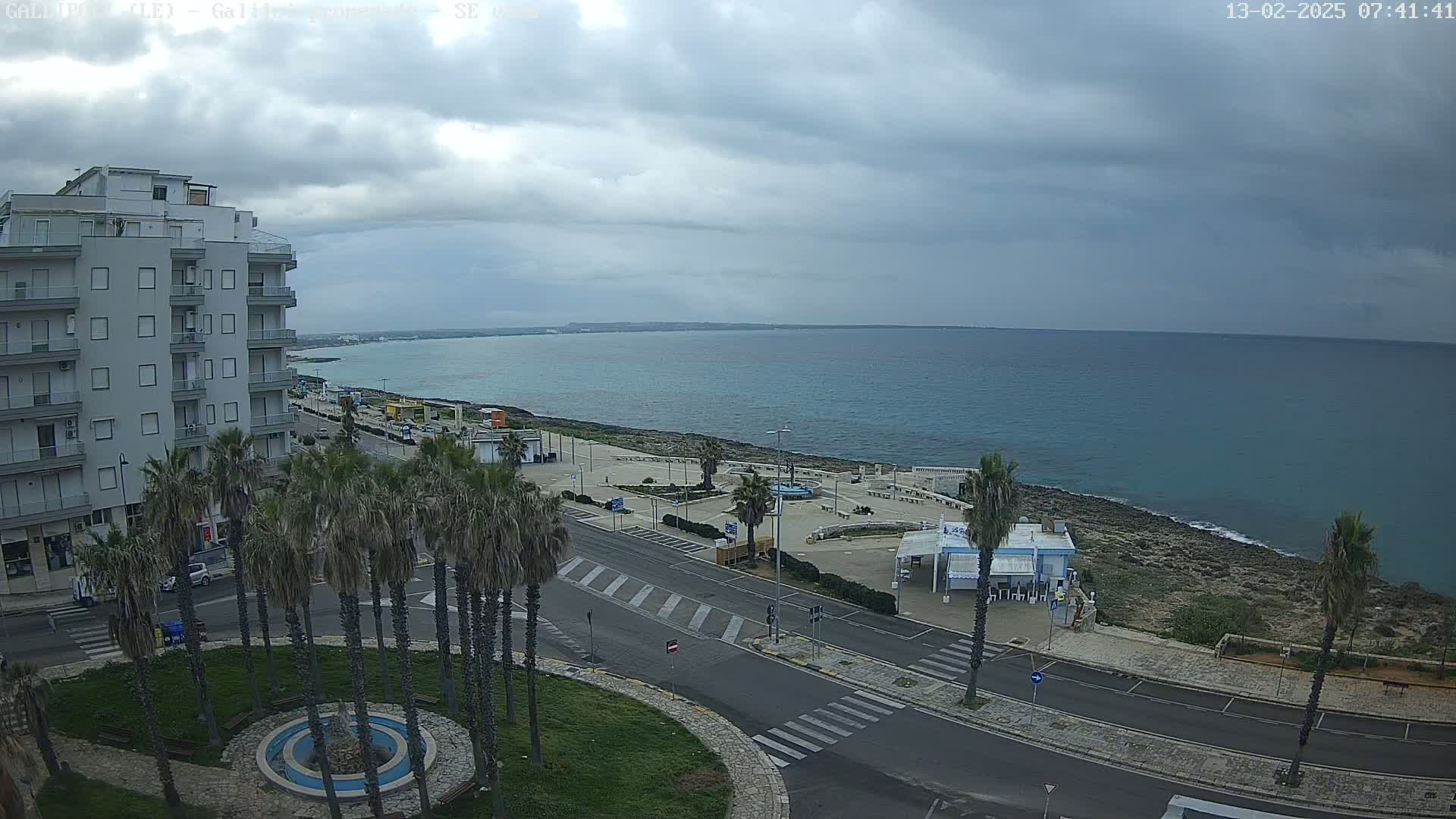 A coastal town scene under a cloudy sky shows a multi-story building, palm trees lining a road leading to a calm sea, and a paved area with some small structures near the shore.