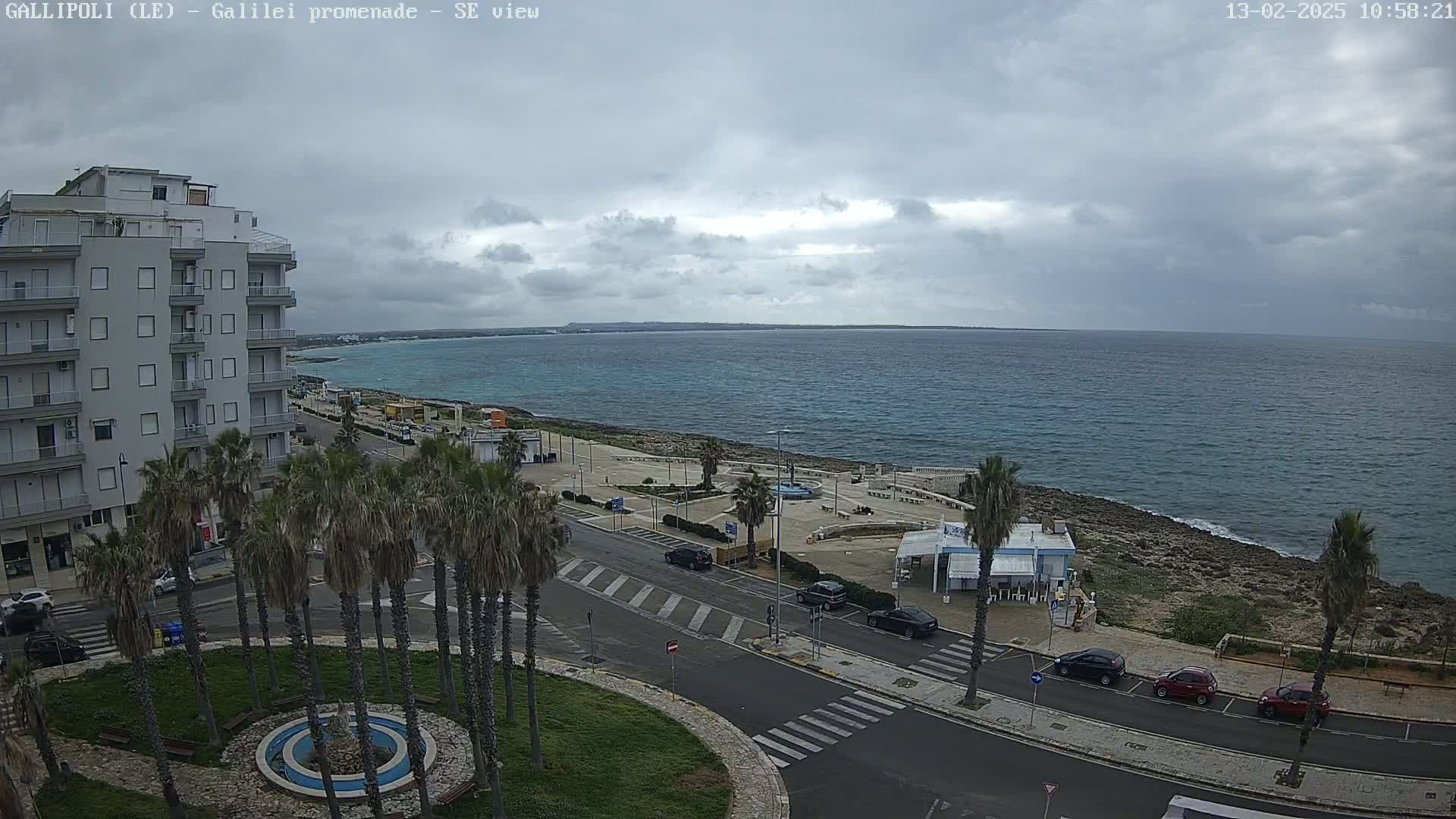 A coastal scene on an overcast day shows a multi-story building, palm trees lining a road next to a promenade overlooking a calm sea.