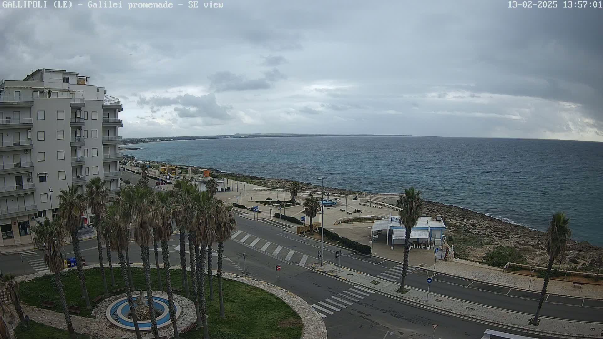 A coastal scene under overcast skies shows a multi-story building, palm trees lining a promenade, and a calm sea extending to a distant horizon.