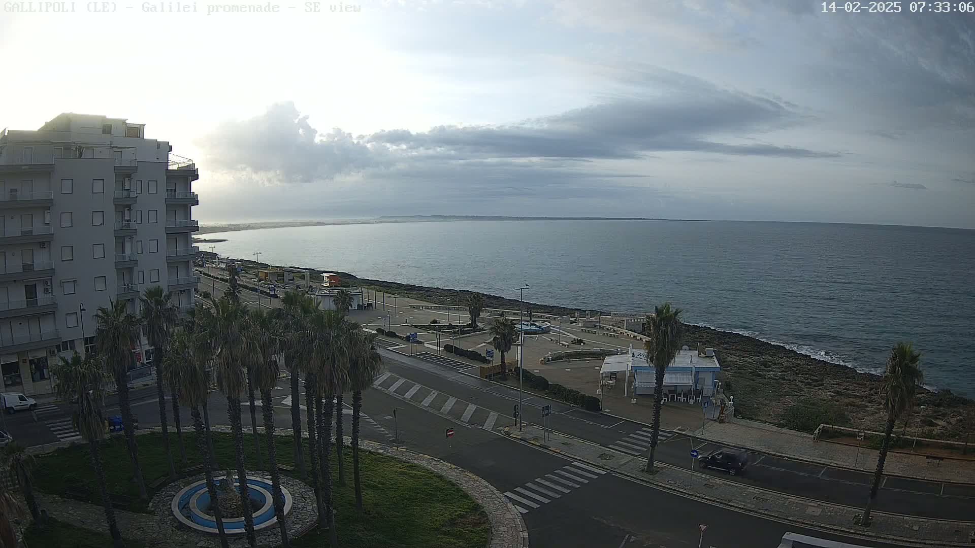 A seaside town scene under a mostly cloudy sky shows a multi-story building, palm trees lining a road leading to a paved seaside promenade, and a calm sea extending to the horizon.