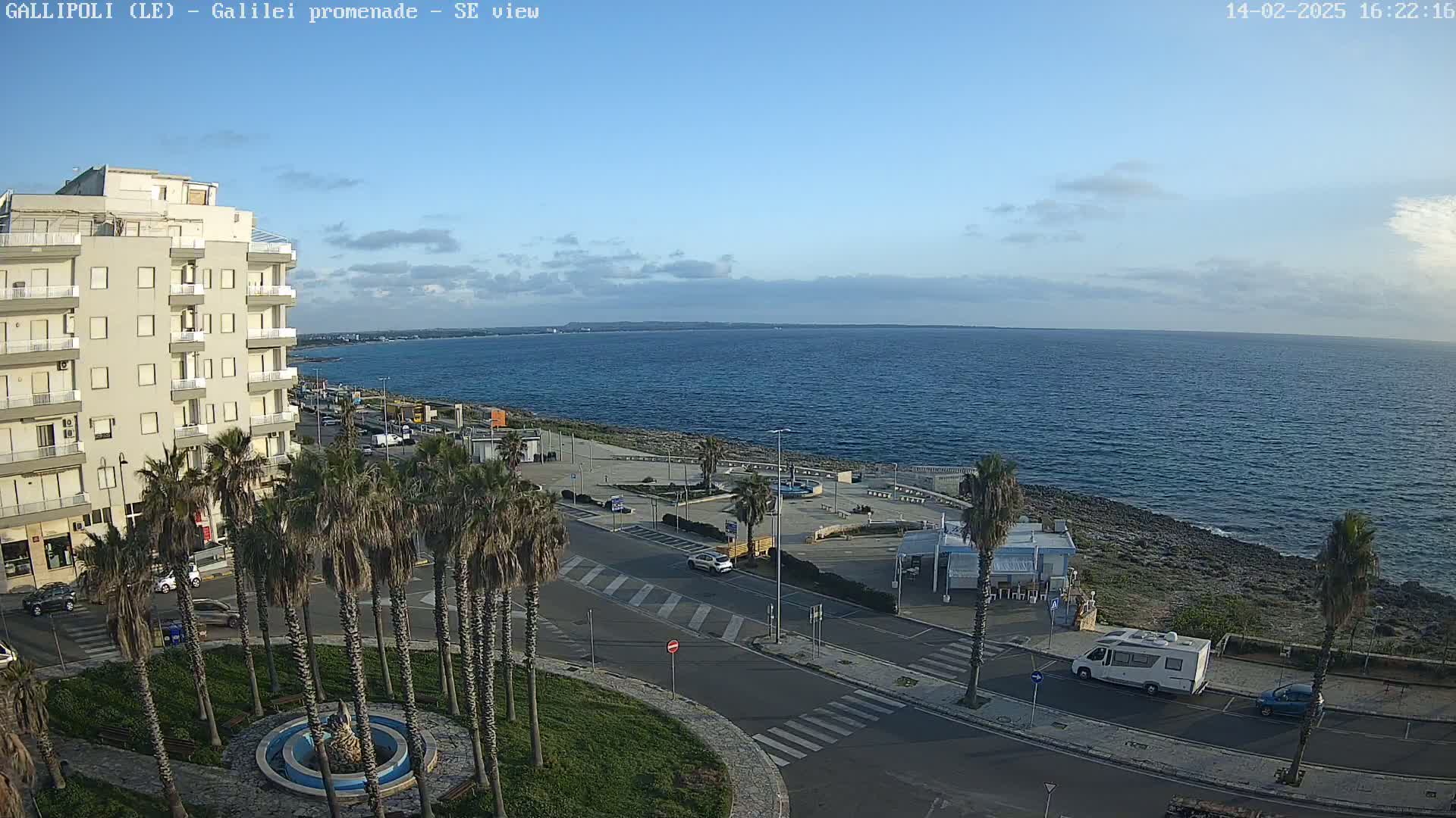 A seaside promenade with palm trees, a multi-story building, and parked cars is visible under a mostly clear sky, with a calm sea extending to the horizon.
