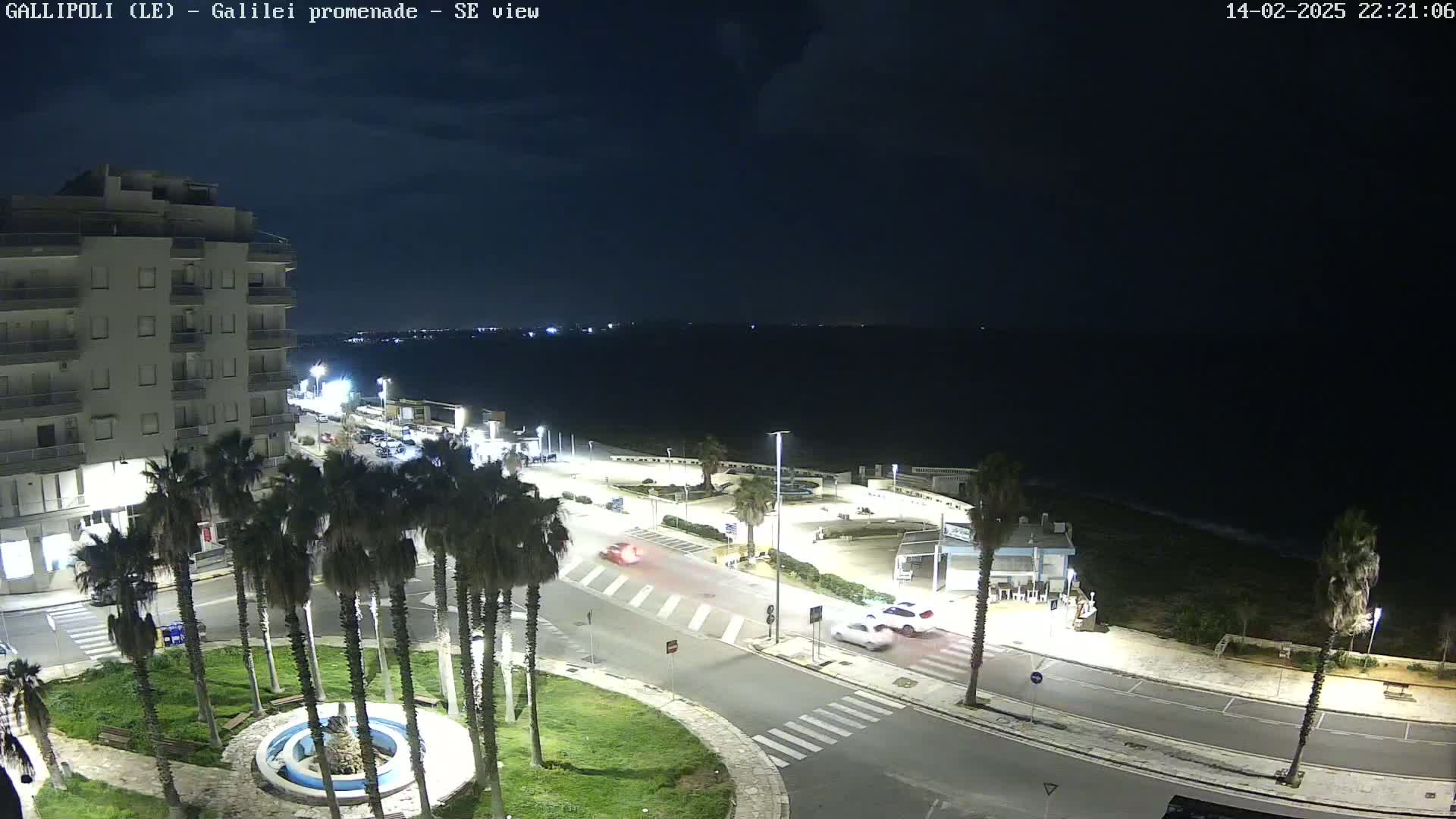A nighttime, high-angle view shows a seaside promenade with palm trees, a multi-story building, a few cars, and a calm, dark sea in the background.