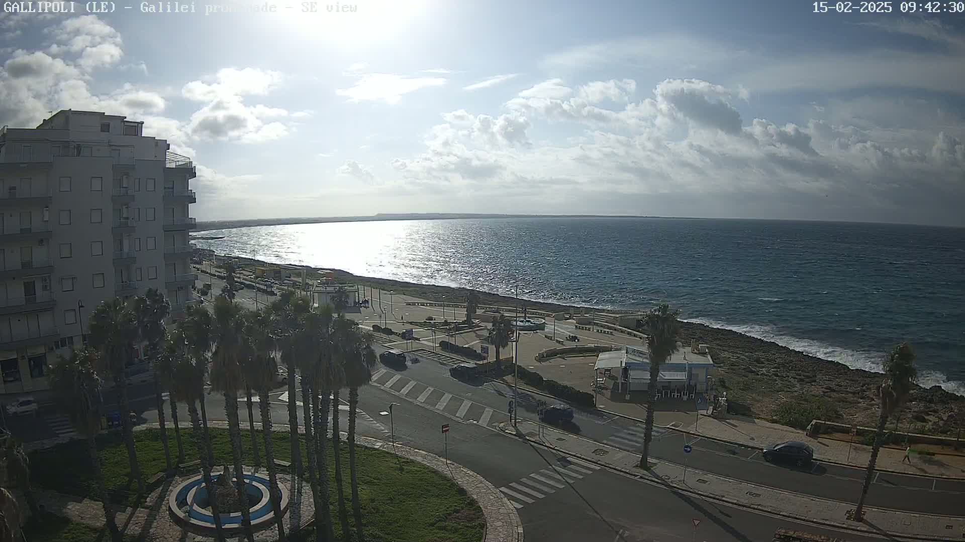 A partly cloudy day reveals a seaside town with a multi-story building, palm trees, a roadway, and a rocky coastline with a calm sea.