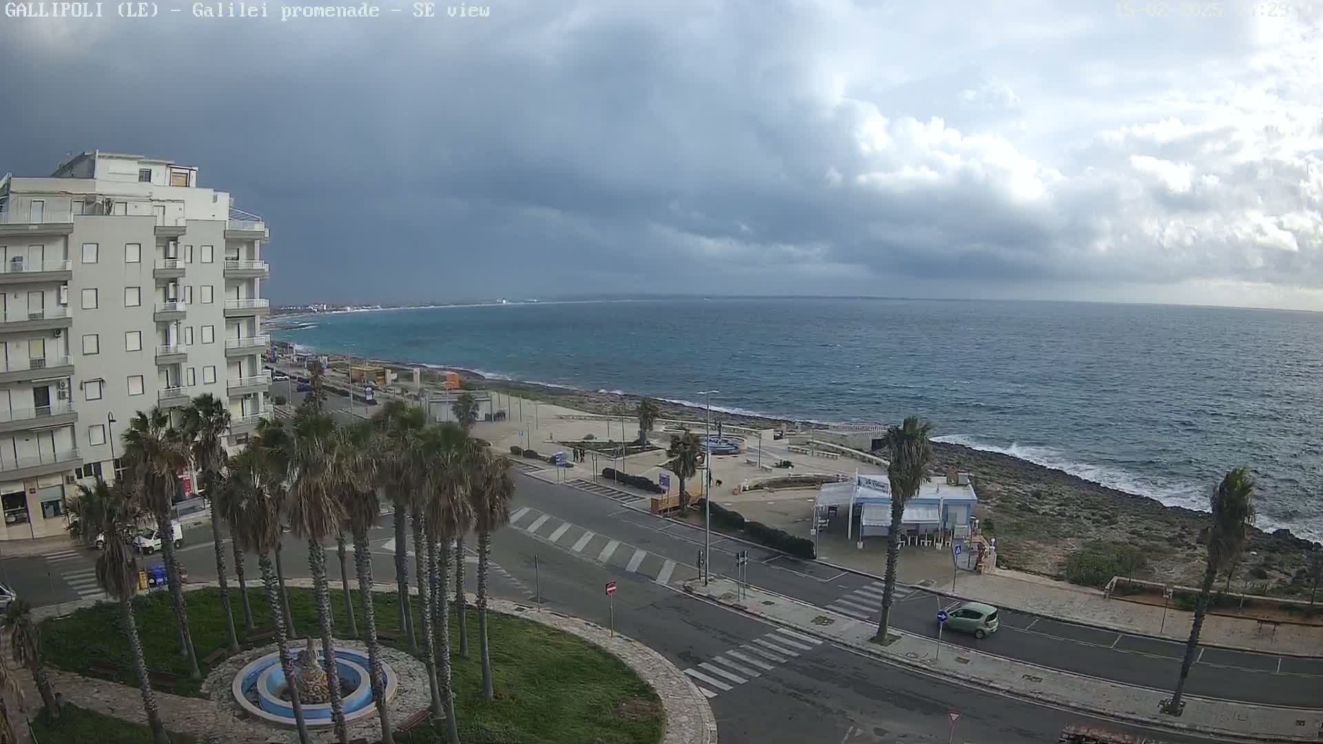 A coastal town scene under a cloudy sky shows a roadway lined with palm trees leading to a rocky shoreline and a calm sea.