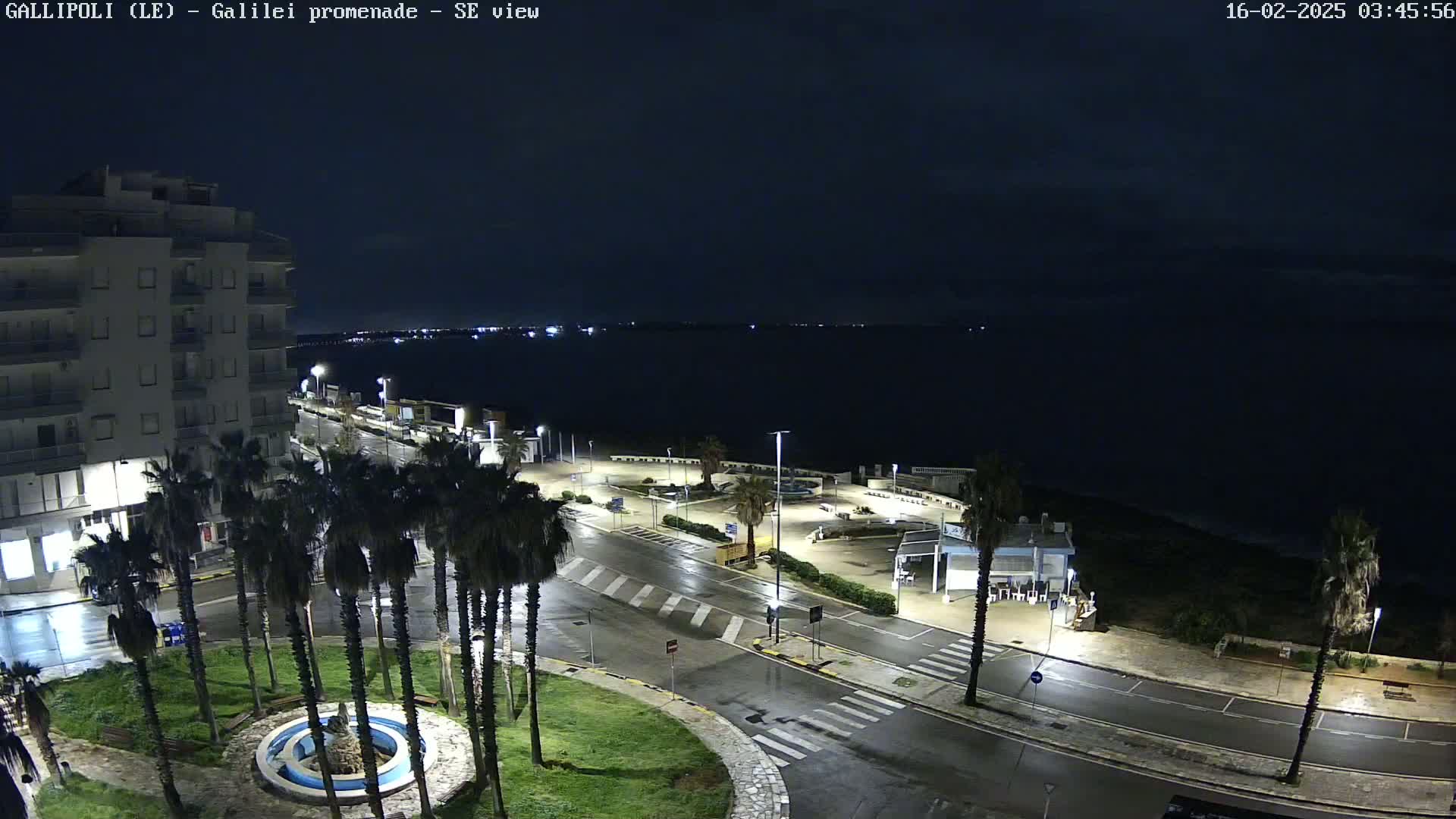 A nighttime, rain-slicked seaside promenade scene shows a multi-story building, palm trees, and a calm, dark sea under a cloudy sky.
