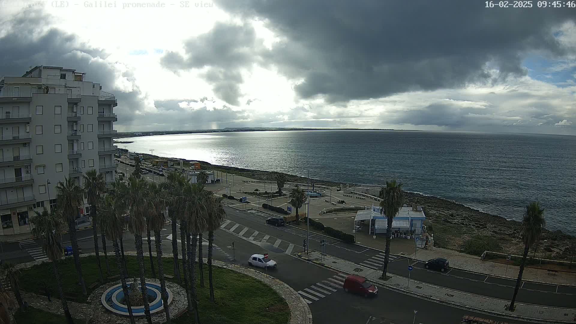 A coastal city scene under a mostly cloudy sky shows a calm sea, a promenade with palm trees and a few cars, and a multi-story building.