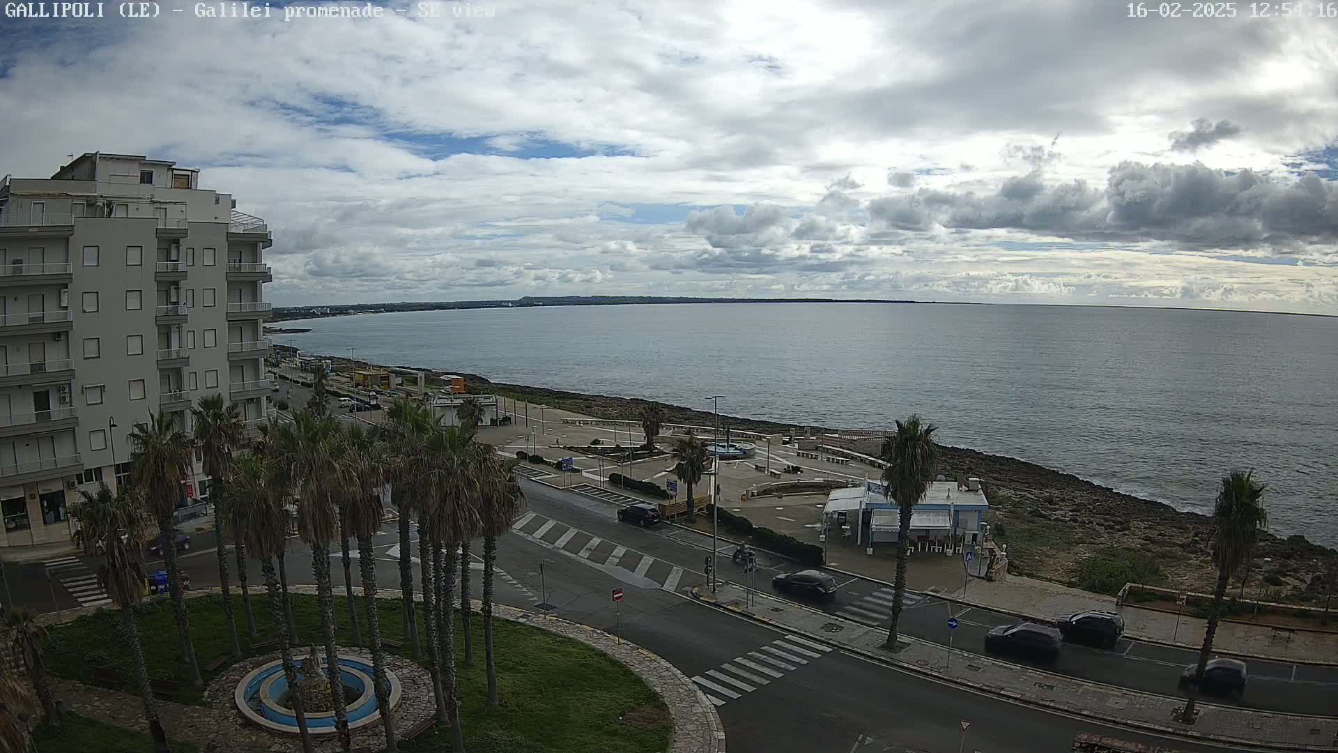 A partly cloudy day overlooks a coastal town with palm trees lining a road next to a calm sea.