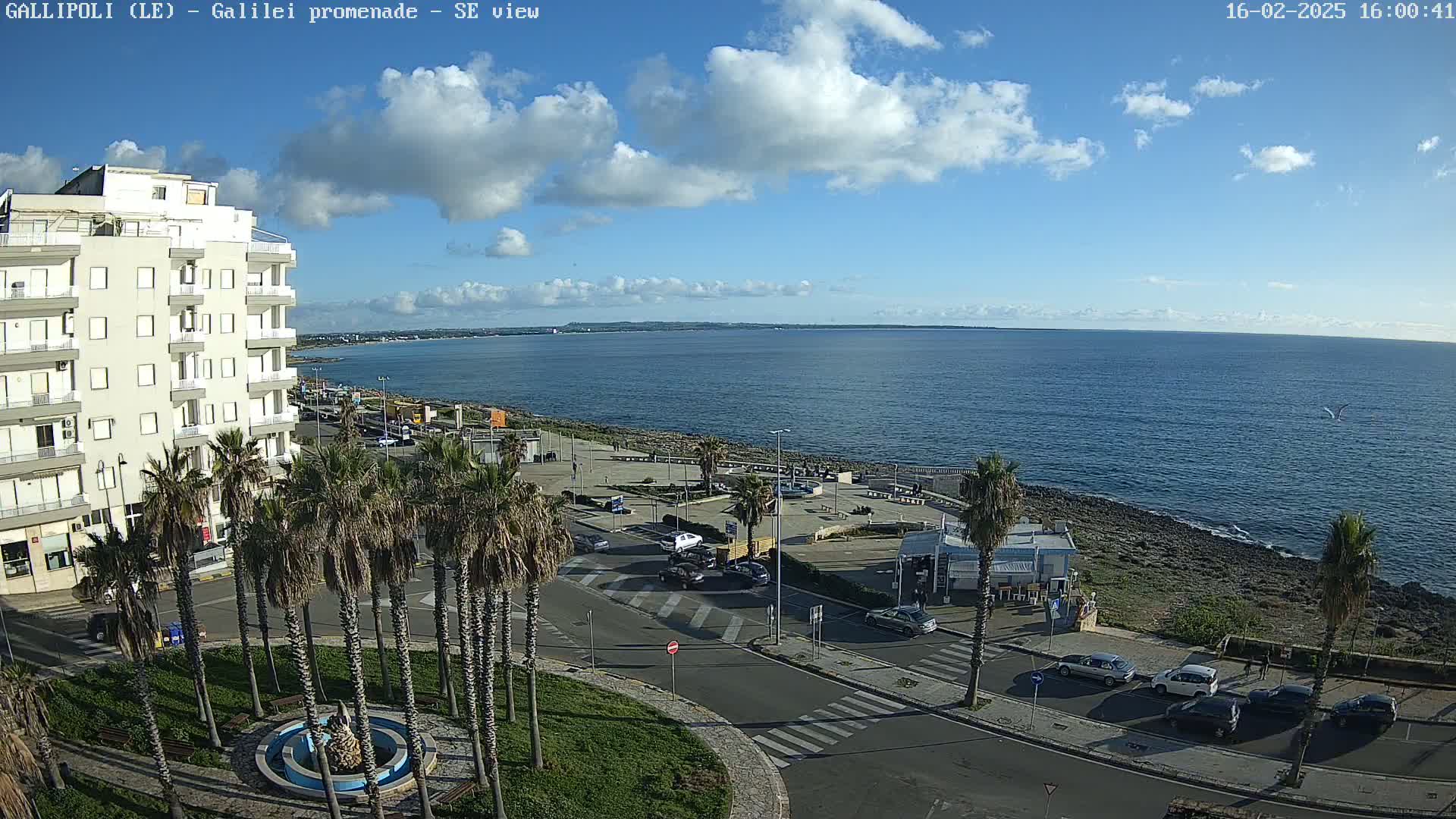 A partly cloudy day overlooks a coastal town square with palm trees, a fountain, and a multi-story building near the ocean.