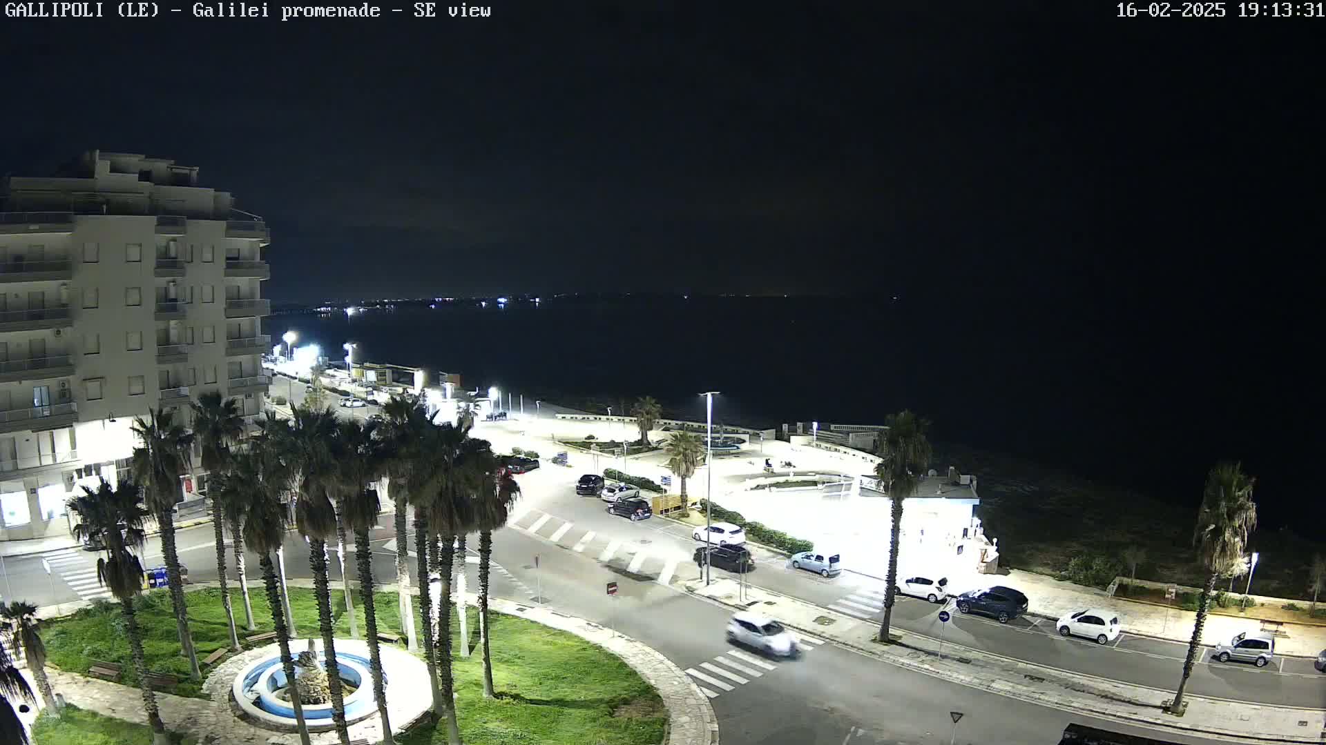 A nighttime, aerial view of a coastal city with a promenade, palm trees, cars parked along a road, and a multi-story building near a calm, dark sea.
