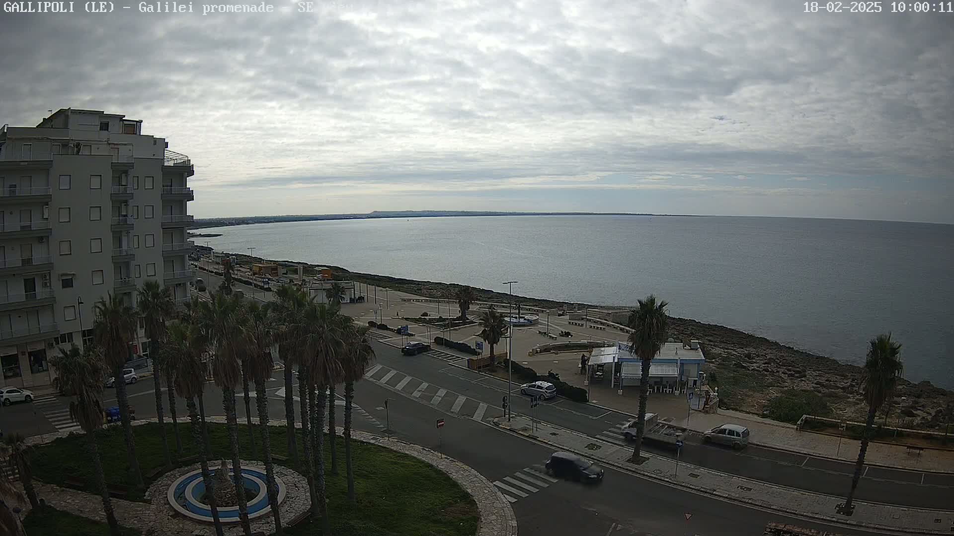 A coastal cityscape under a cloudy sky shows a multi-story building, palm trees, a road, a promenade along a rocky shoreline, and a calm sea extending to the horizon.