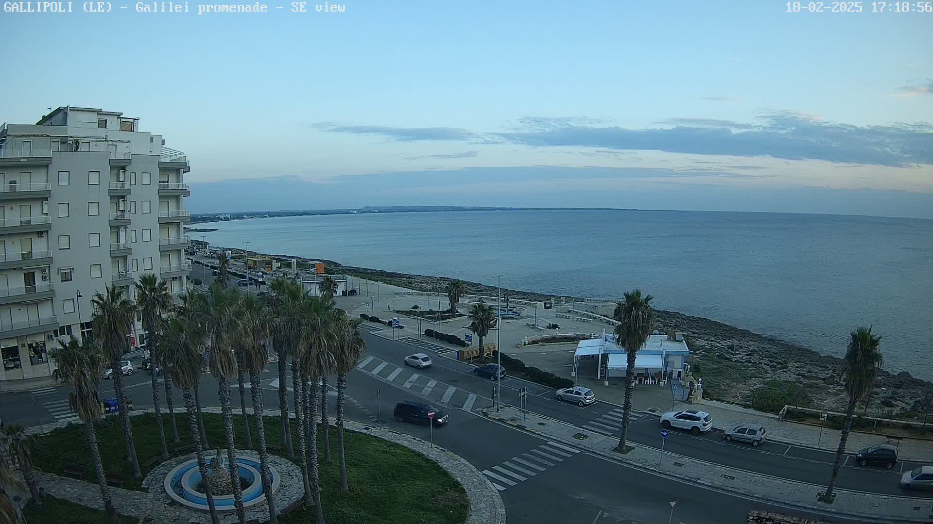 A seaside promenade with palm trees, cars, and a multi-story building overlooks a calm sea under a mostly clear, twilight sky.