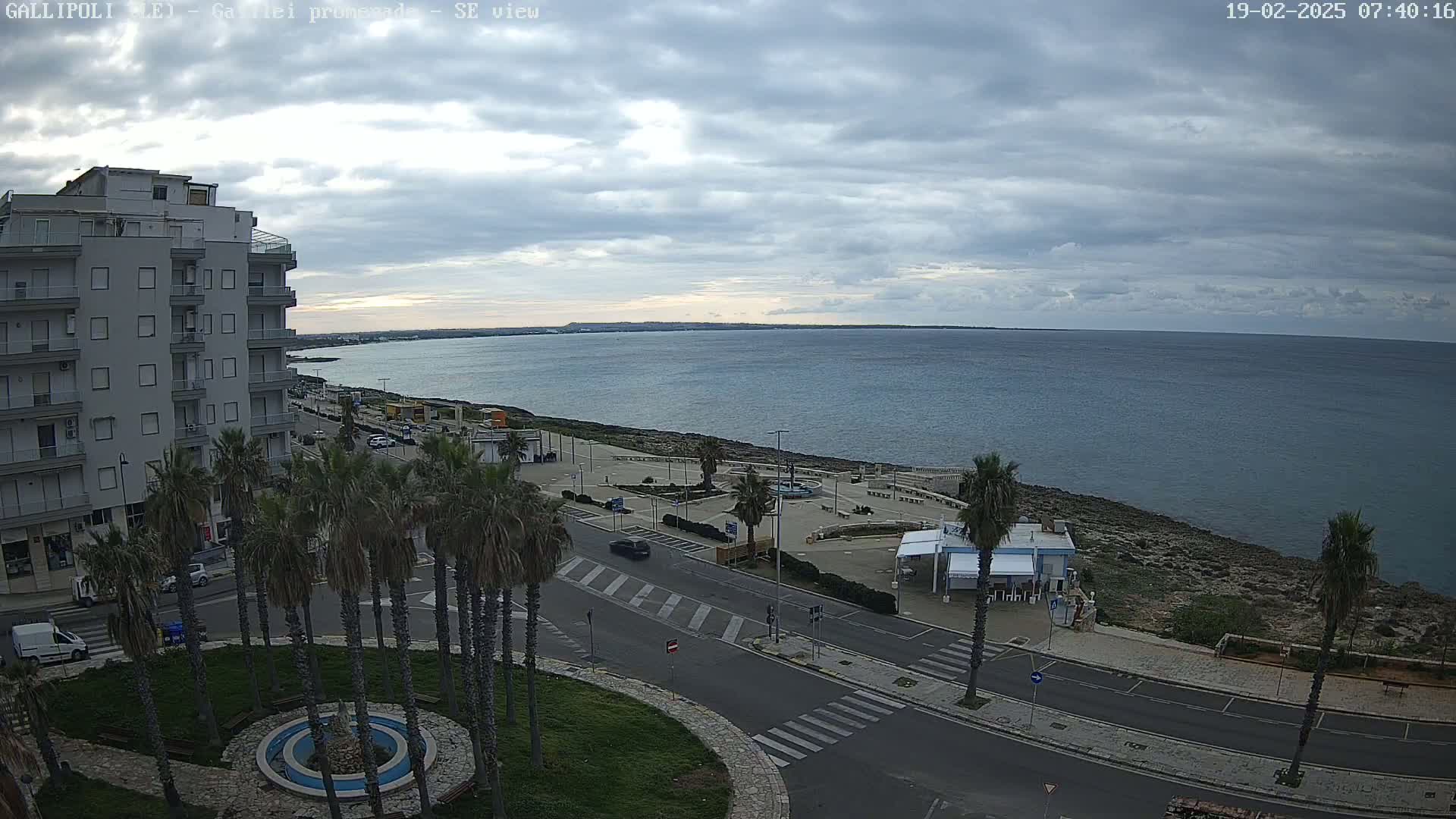 A mostly cloudy day overlooks a coastal town with palm trees lining a street next to a calm sea.