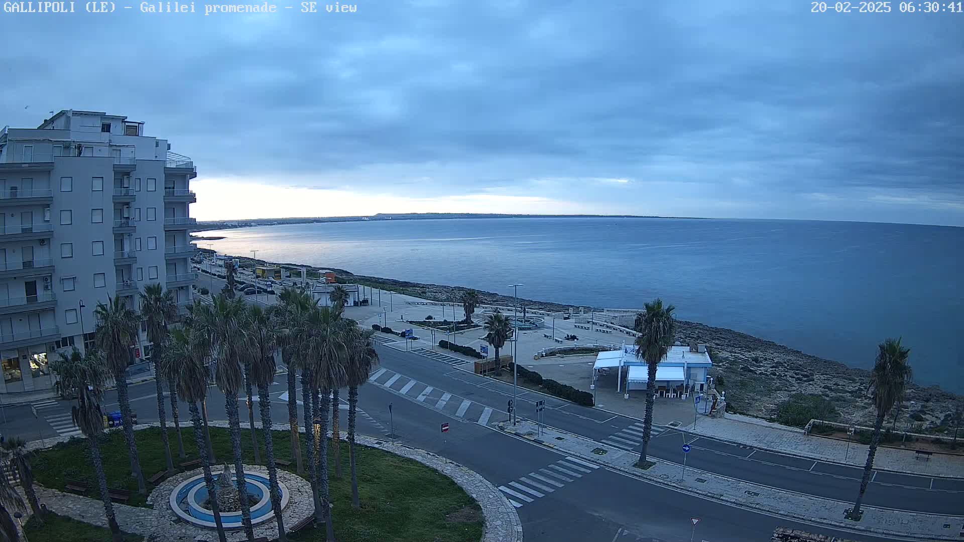A seaside promenade with palm trees and a multi-story building is visible under a mostly cloudy sky.