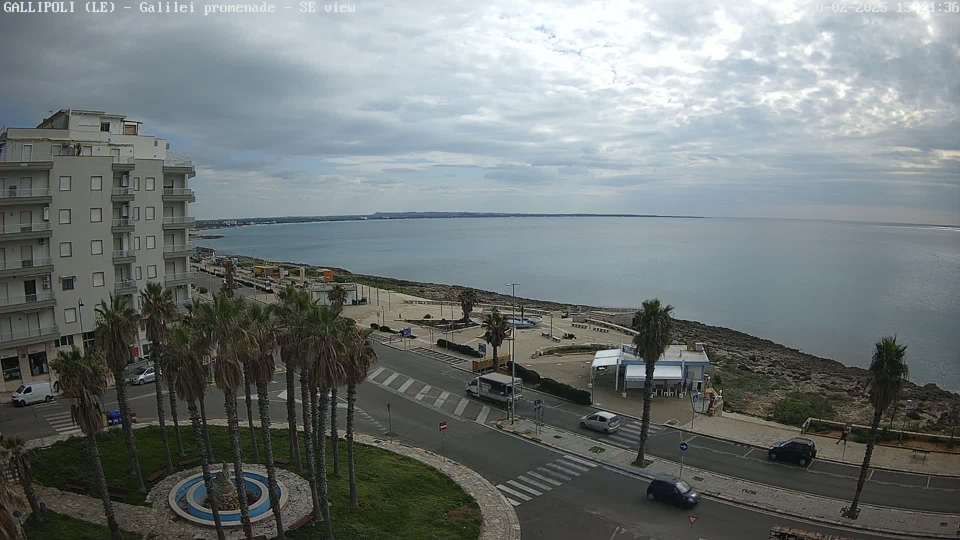 A seaside promenade with palm trees, buildings, and a calm sea under a cloudy sky.