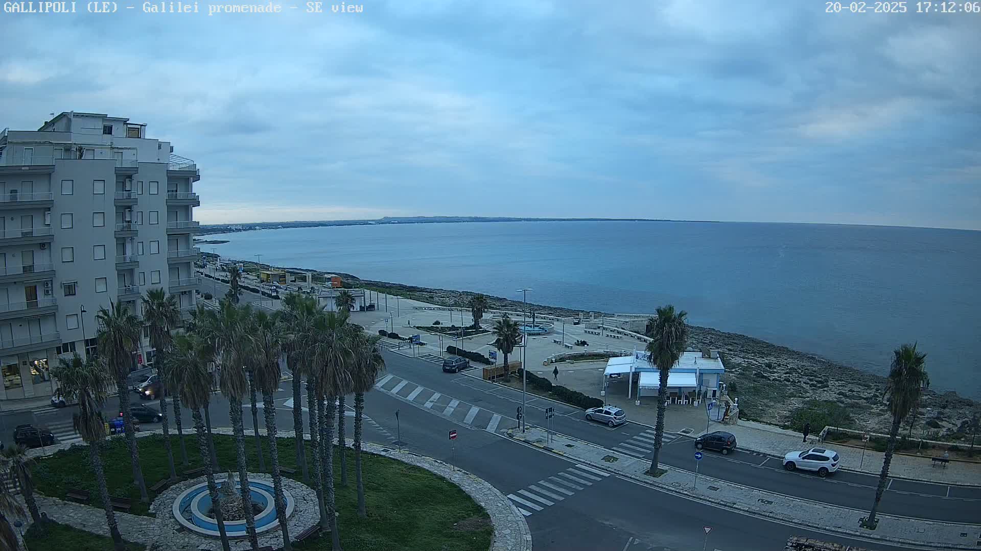 A seaside town scene under a partly cloudy sky shows a calm sea, a paved coastal road with parked cars, palm trees, and a multi-story building.