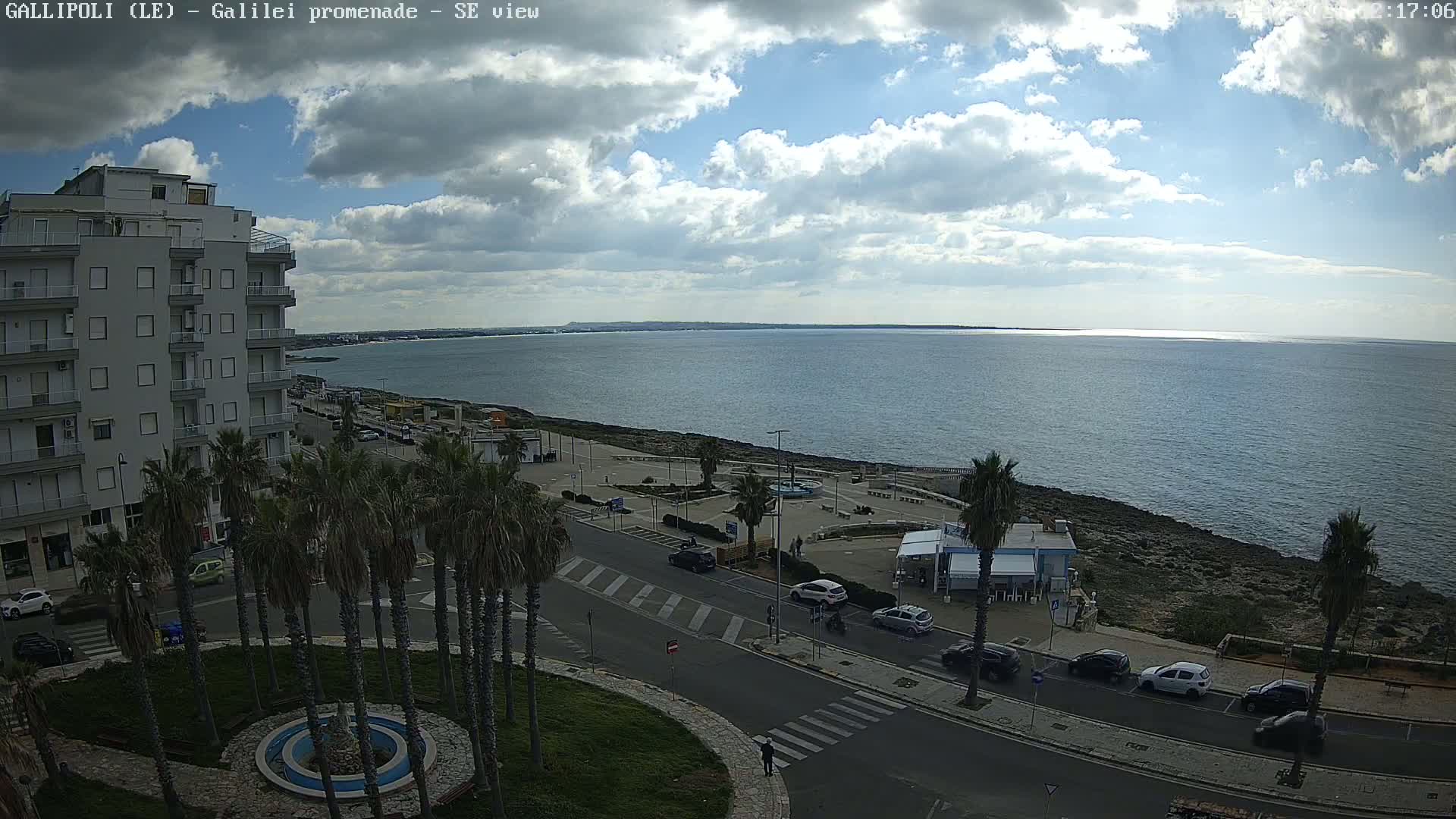 A partly cloudy day shows a seaside promenade with palm trees, parked cars, and a multi-story building overlooking a calm sea.