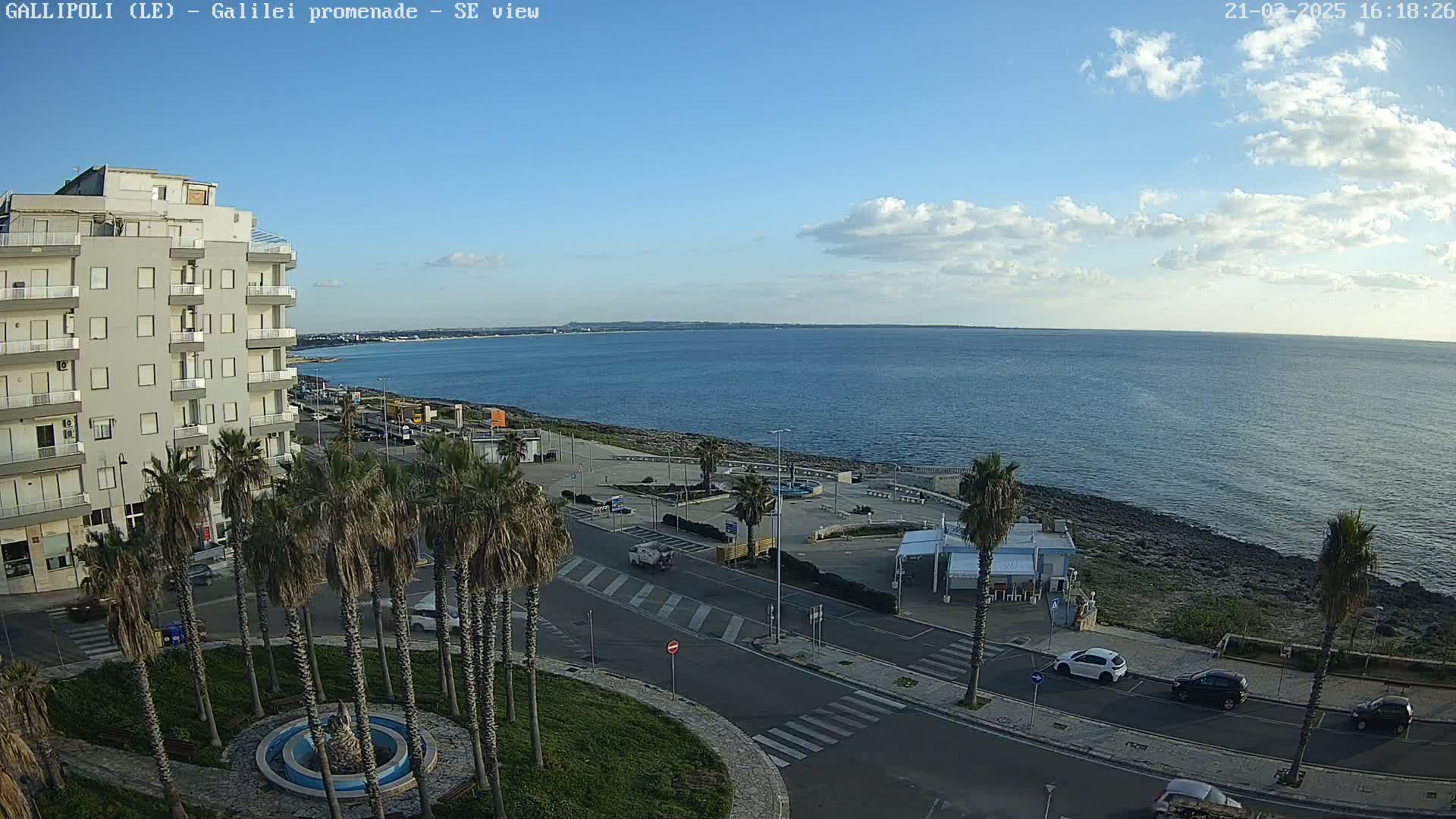 A partly cloudy day shows a seaside promenade with palm trees, a multi-story building, and a few cars parked along the road next to a calm, blue sea.