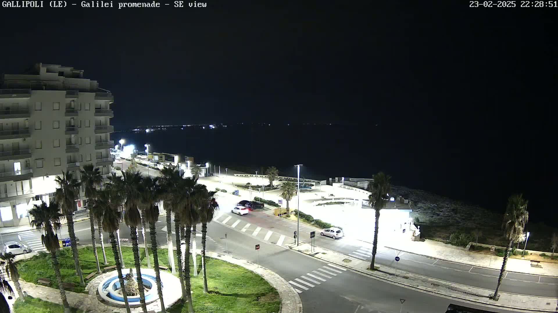 A nighttime, high-angle view shows a coastal town with palm trees lining a street next to a calm sea under a dark sky.