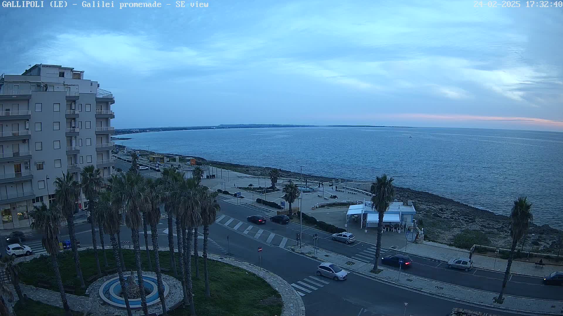 A seaside promenade at dusk shows a calm sea under a mostly clear, dusky blue sky, with palm trees lining a road beside a building and a small plaza.