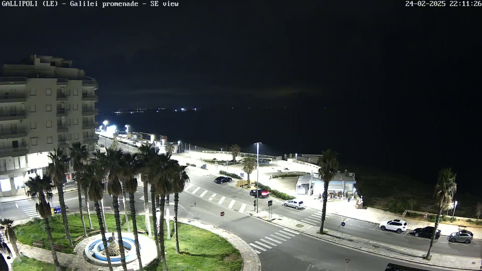 A nighttime view of a coastal town square with palm trees, a building, cars parked along roads, and a dark, calm sea in the background.