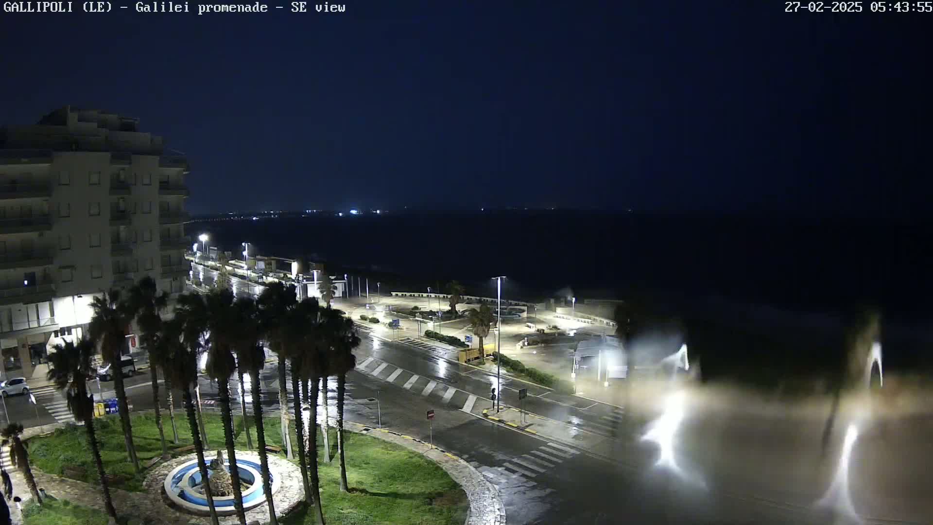 A nighttime, rain-soaked coastal scene shows a multi-story building, palm trees, and wet roads near a dark ocean.