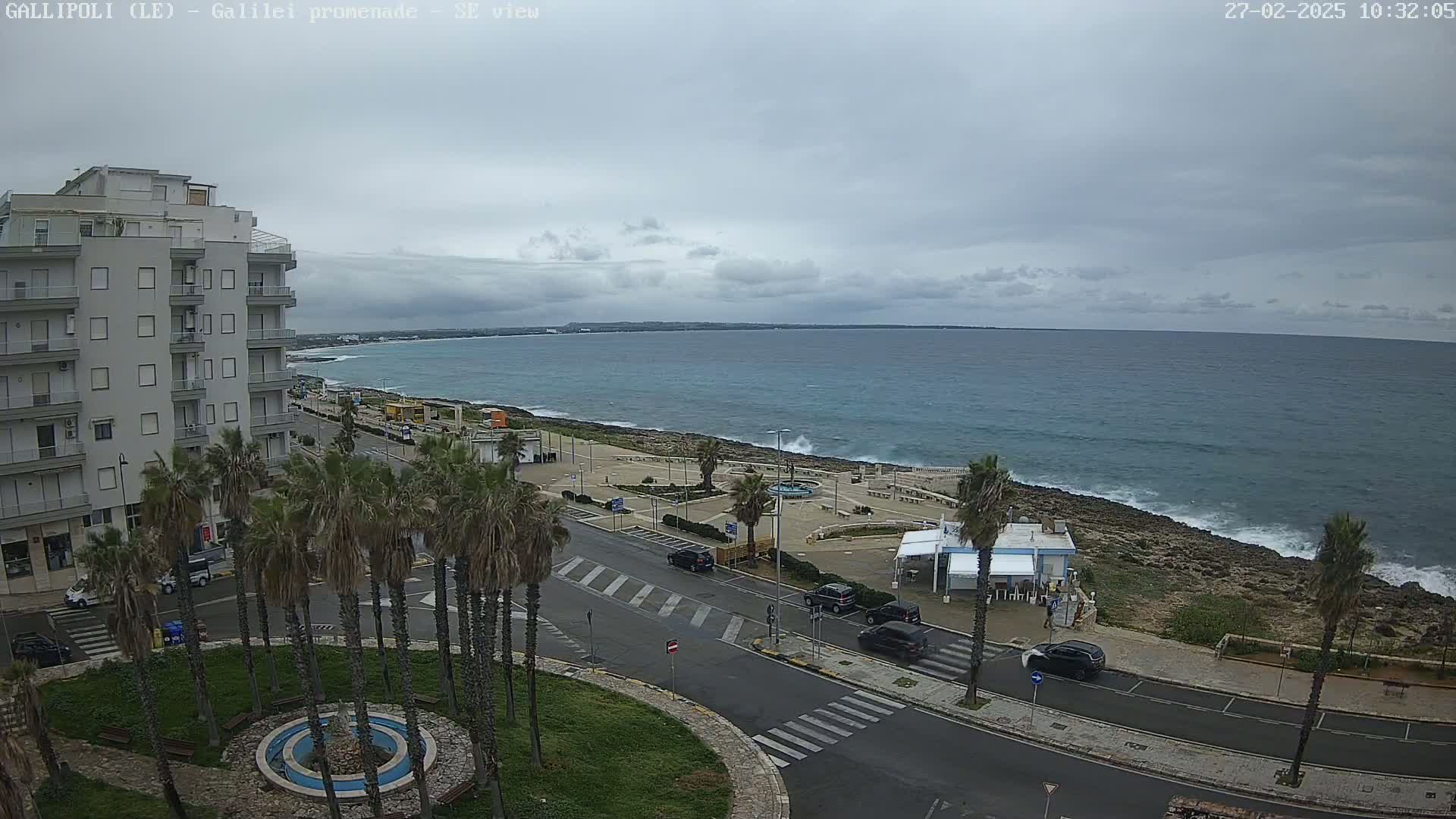 A coastal road lined with palm trees runs alongside a rocky shoreline with a partly cloudy sky overhead, and a calm sea with small waves.