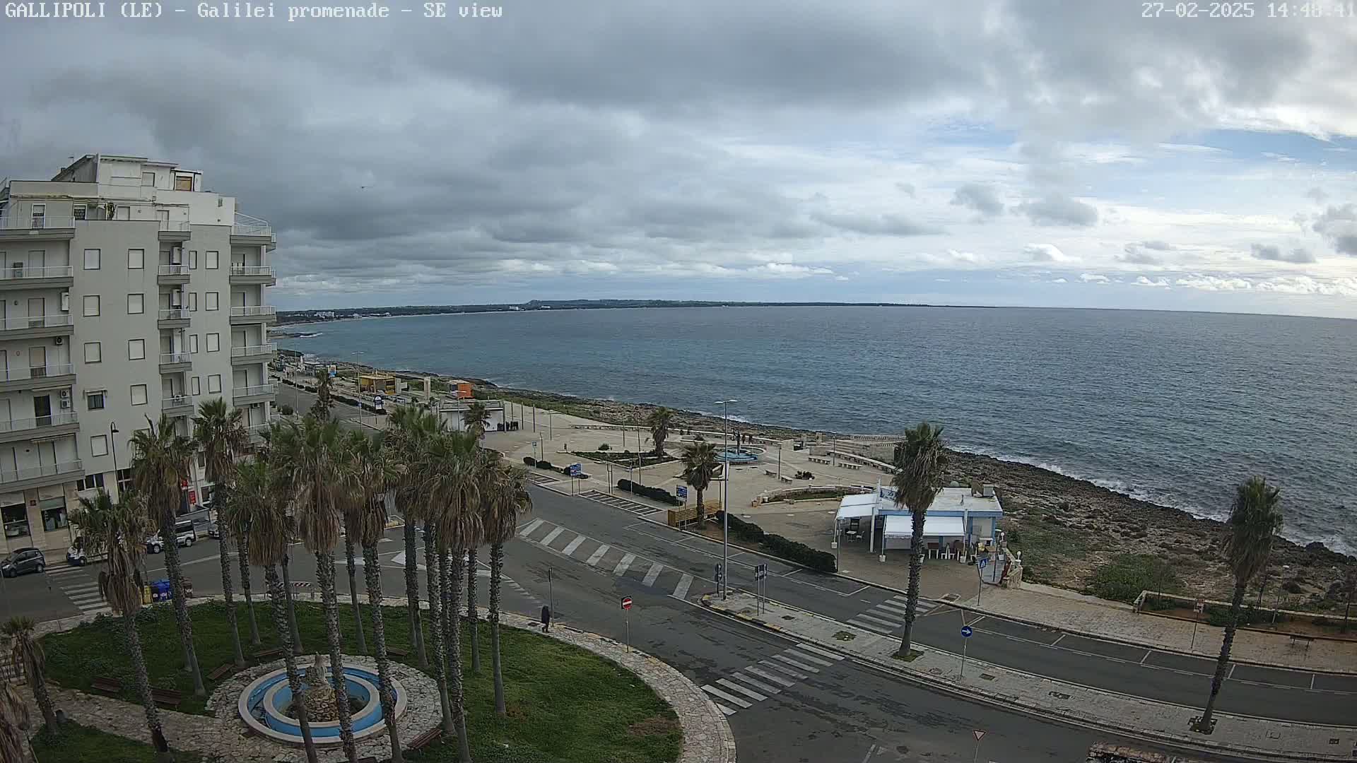 A seaside promenade with palm trees, a multi-story building, and a calm sea under a cloudy sky.