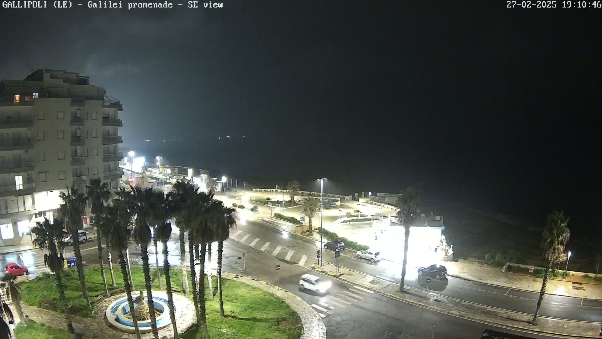 A nighttime, rain-soaked coastal scene shows a multi-story building, palm trees lining a wet roadway, and vehicles driving through a town square overlooking a dark sea.