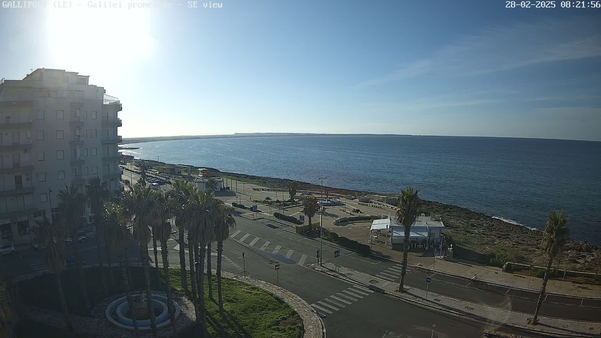 A high-angle, sunny view shows a calm ocean stretching to a distant shoreline, with a coastal road lined with palm trees leading to a building, and a plaza with a fountain in the foreground.